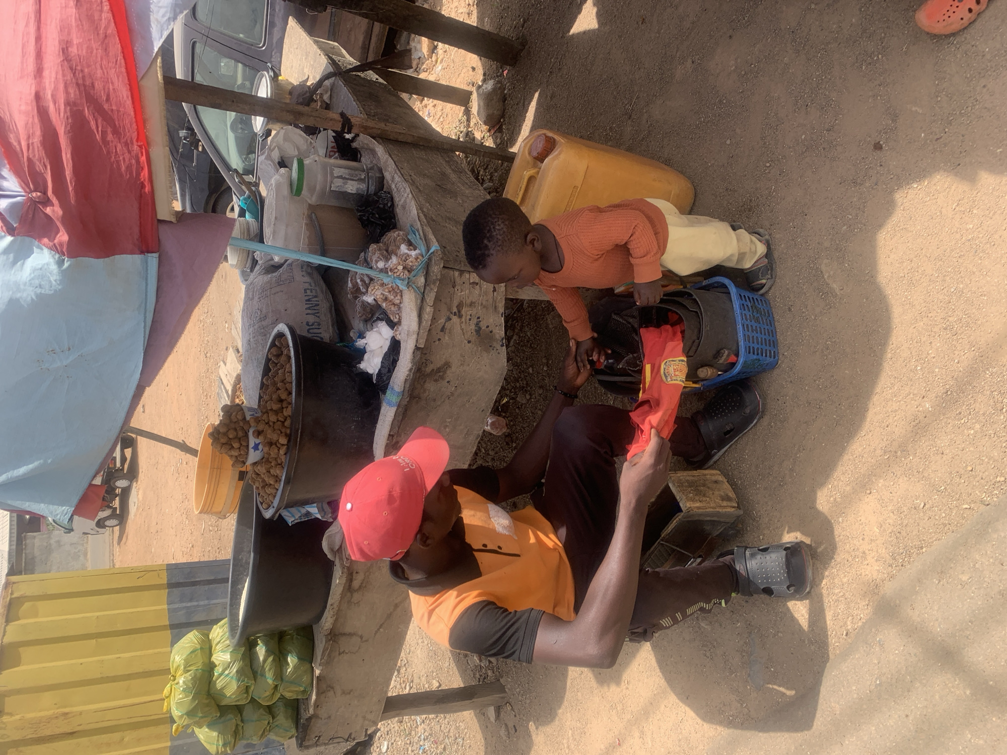 This outdoor image captures a market scene in what appears to be Jos, Nigeria, during a bright, sunny day, indicated by the strong shadows cast on the dusty ground.
The central figures are an adult male and a young child. The man, wearing an orange t-shirt over a black long-sleeved top, black trousers, and a red cap, is seated on a low structure, possibly part of a stall. He wears black sandals and is holding a piece of red fabric in his hands, possibly merchandise. Adjacent to him, a young child, dressed in an orange long-sleeved shirt, light-colored trousers, and dark shoes, stands and looks down into a blue plastic basket near the man.
The scene is set around a makeshift market stall constructed from wooden planks and possibly concrete, covered by a colorful canopy of red, light blue, and white fabric. The stall is laden with various goods. Large black basins hold what appear to be groundnuts (peanuts) and another staple like grains or flour. Yellow buckets and a large yellow jerry can are also visible on the ground. Several items are packed into green plastic bags, and a white sack with the partial text "PENNY SU" is also present. In the background, a dark-colored car is partially visible, parked near the stall. An orange sandal, similar in style to the man's black sandals, is visible in the top right corner, suggesting another person nearby. The overall impression is a bustling, informal market environment where daily commerce takes place.