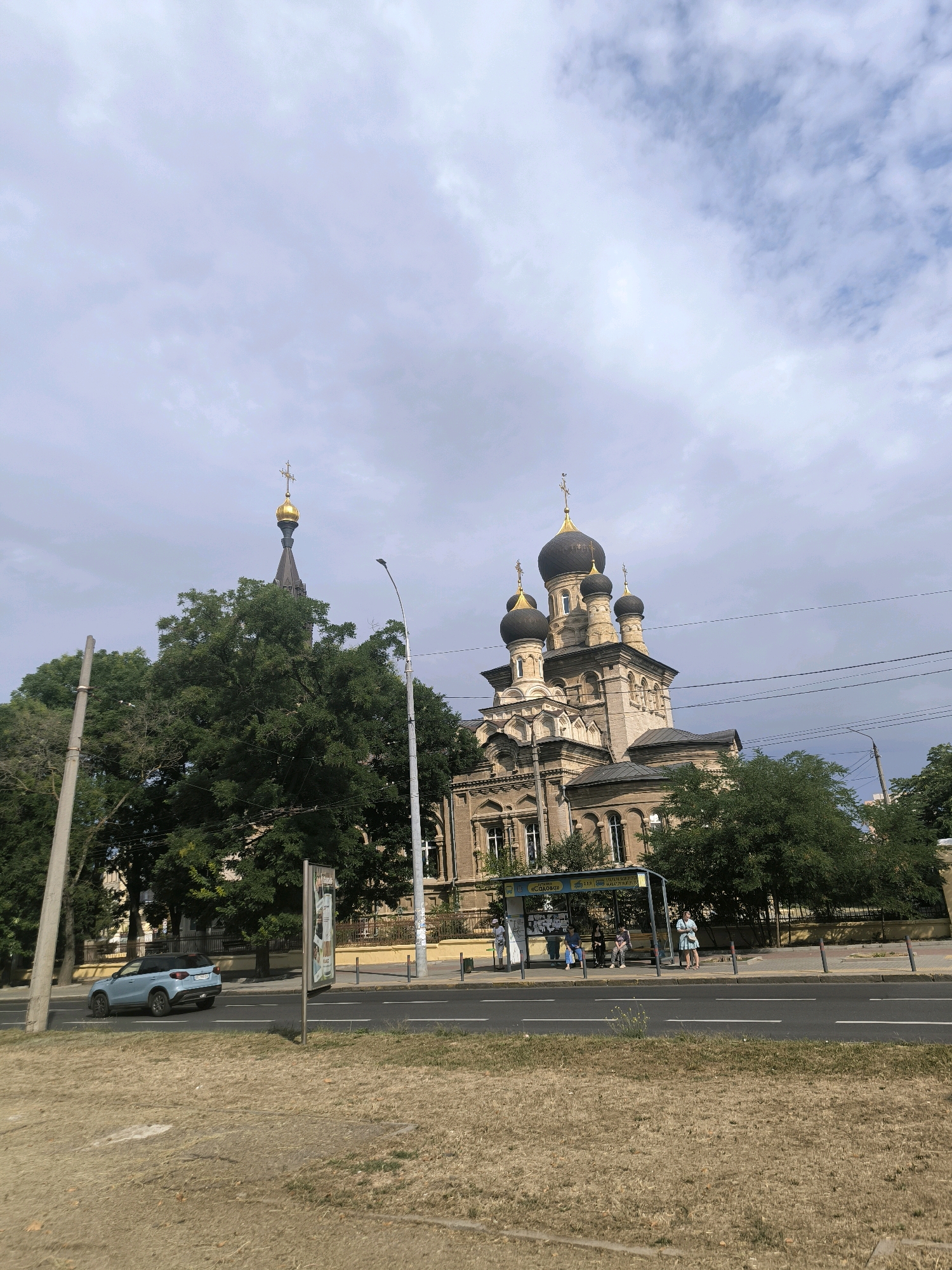 The image depicts a street scene in Mykolaiv, Ukraine, featuring a prominent Orthodox church with multiple domes and a golden cross atop its highest spire. The church is characterized by its intricate architectural details and light-colored brickwork. Adjacent to the church, a bus stop shelters several individuals, suggesting public transportation usage. A light blue car is observed driving on the asphalt road, which is marked with white lane lines. Lush green trees are interspersed throughout the scene, providing a contrast to the urban structures. The sky is partly cloudy, with a mix of gray and white formations. The foreground shows a dry, grassy area.