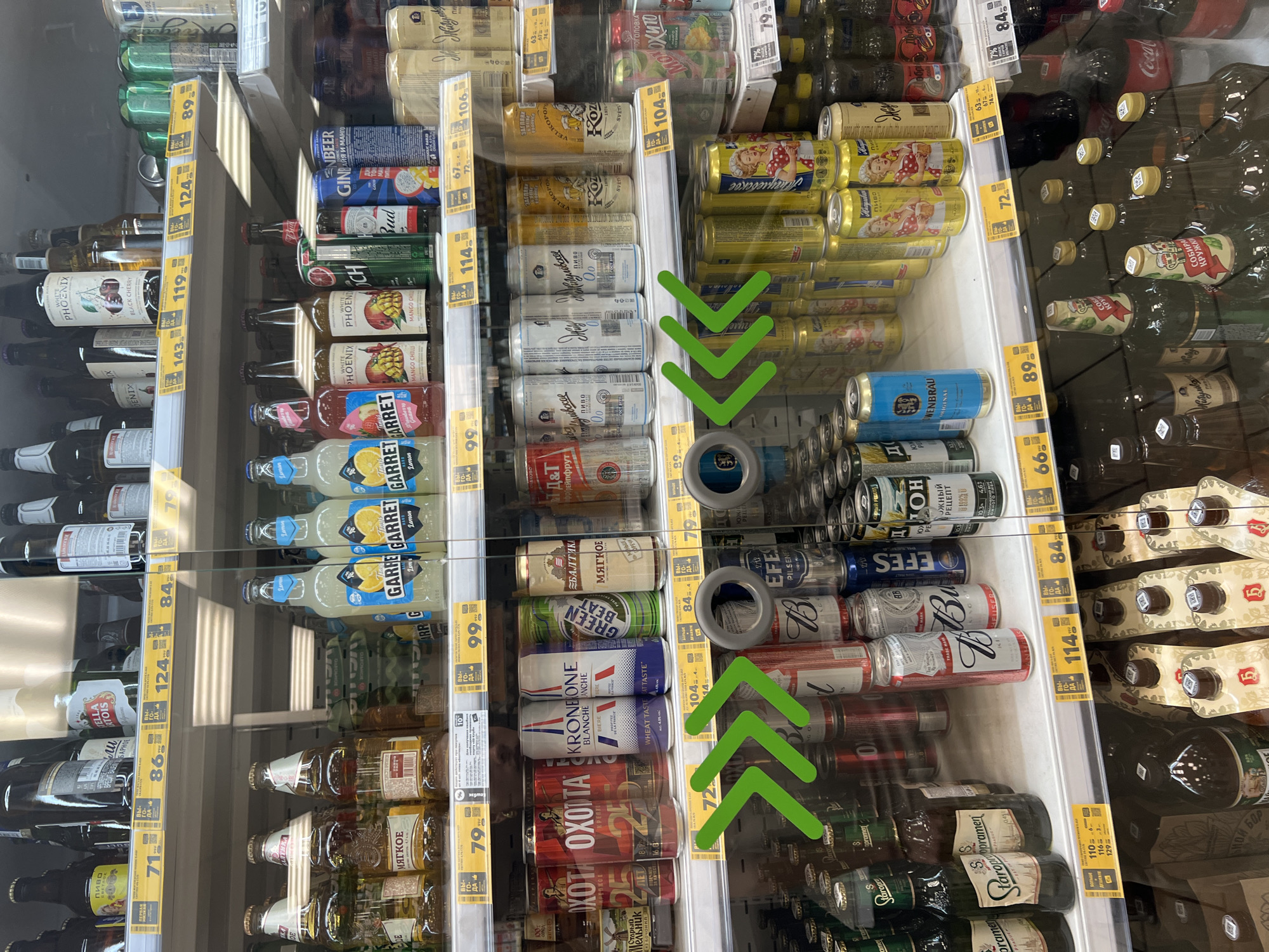 This is a photograph of the interior of a grocery store, likely a supermarket, focusing on a refrigerated display case filled with beverages. The display is divided into several shelves, each stocked with a variety of canned and bottled drinks.
The primary subjects are the beverages themselves, predominantly beer and possibly other alcoholic or non-alcoholic drinks. The cans are arranged in rows, with brands and labels visible, though many are partially obscured. Some bottles, particularly at the bottom and along the sides, are also present. The overall impression is one of abundance and variety.
The scene is indoors, in a commercial setting. The lighting appears to be artificial, typical of a store environment, with some reflections visible on the glass of the display case. The setting suggests a self-service retail environment where customers can select their preferred drinks.
Notable details include the presence of price tags or promotional labels on the shelves, some with Cyrillic text. Several green checkmarks, resembling a "skip" or "selection" symbol, have been superimposed onto the image on the right side, indicating a possible selection or emphasis of certain items or areas within the display. The text visible is predominantly in Russian, with brand names like "Bud," "GİN," "KRONONE," "OXOTA," "GARRET," "WHITE PHOENIX," "Staropramen," "EFES," and "Baltika" being identifiable. Other text includes pricing information and product descriptions in Russian. There are no people visible in the frame. The time of day and weather are not discernible from the image. The location context of Krasnodar, Russia, is provided, which aligns with the visible Russian text.