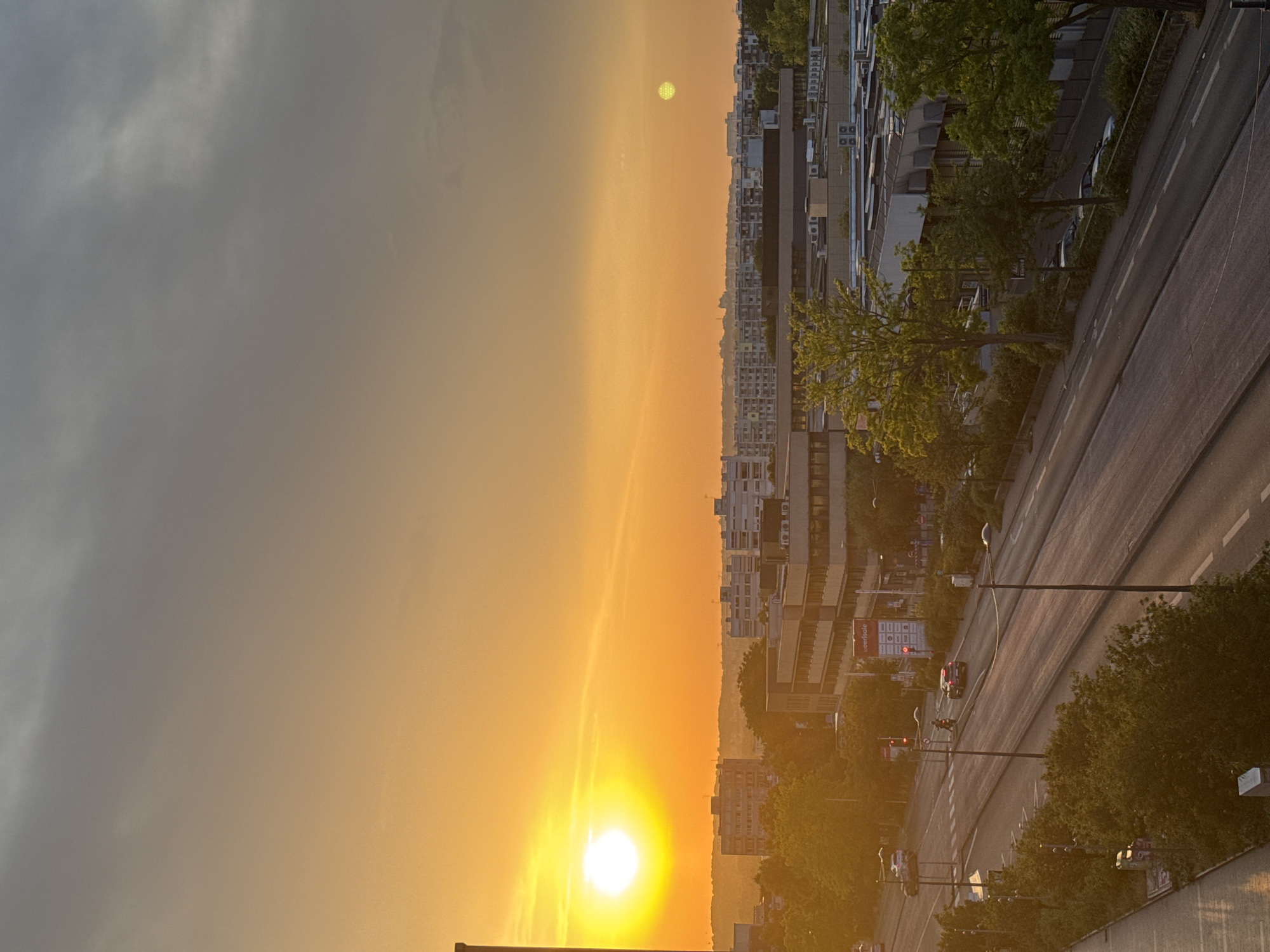 This is a wide shot from an elevated vantage point looking over a city street at sunset. The sky is a gradient of orange and yellow near the horizon, transitioning to a muted gray at the top, with some clouds visible. A bright, white sun is prominently displayed on the left side of the frame, casting a warm glow over the scene. In the distance, a range of city buildings, including apartment blocks and commercial structures, are visible. Trees are scattered throughout the cityscape, some with vibrant green foliage catching the sunlight.
A two-lane road runs diagonally through the lower portion of the image, with vehicles visible, including cars and a motorcycle. Streetlights and traffic signals are present along the road. The sidewalks are lined with trees and bushes, and some parked cars are visible. A sign with "woerlsale" is affixed to one of the buildings. In the upper right corner of the sky, a pale, yellowish-green circular object, possibly a secondary sun or a bright atmospheric phenomenon, is visible. The overall impression is of a peaceful, urban setting bathed in the warm, fading light of sunset.