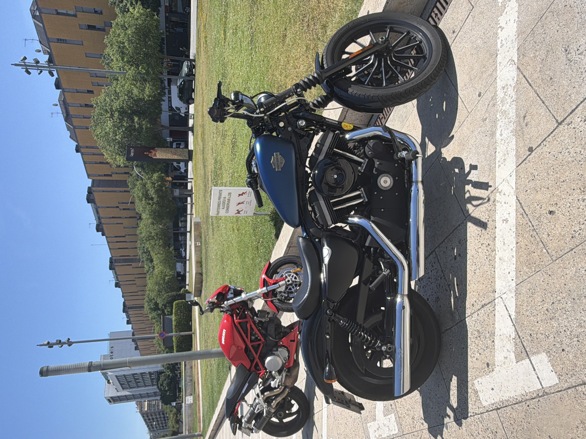 The image shows two motorcycles parked on a paved area in L'Hospitalet de Llobregat, Spain. The motorcycles are a blue Harley-Davidson and a red Ducati. They are parked next to a grassy area and in the background there are buildings and trees. In the background there is a road with vehicles in it. There is a sign near the Harley-Davidson. The lighting suggests it is daytime.