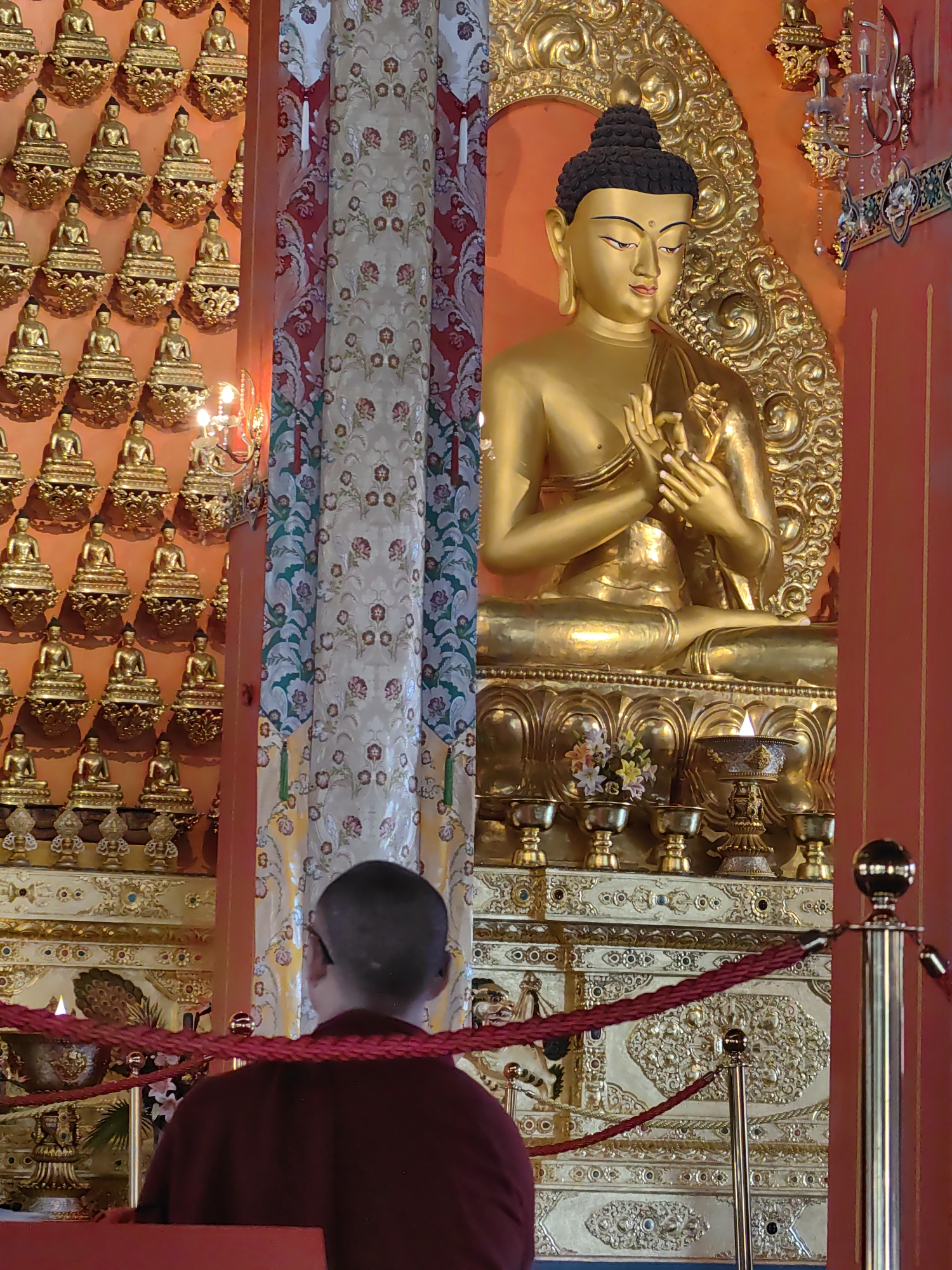In Parsa, Nepal, a monk in a maroon robe faces away from the camera, looking towards a large, ornate golden statue of Buddha. The statue is seated on a tiered pedestal adorned with intricate gold carvings and small floral offerings. Behind the Buddha, the wall is covered in a grid of smaller golden Buddha figurines, each on its own decorative shelf. To the left of the statue, a patterned fabric hangs, featuring a floral design in red, white, and green. Red ropes on stanchions in the foreground create a barrier. The scene is illuminated by soft lighting, casting a warm glow on the golden figures.