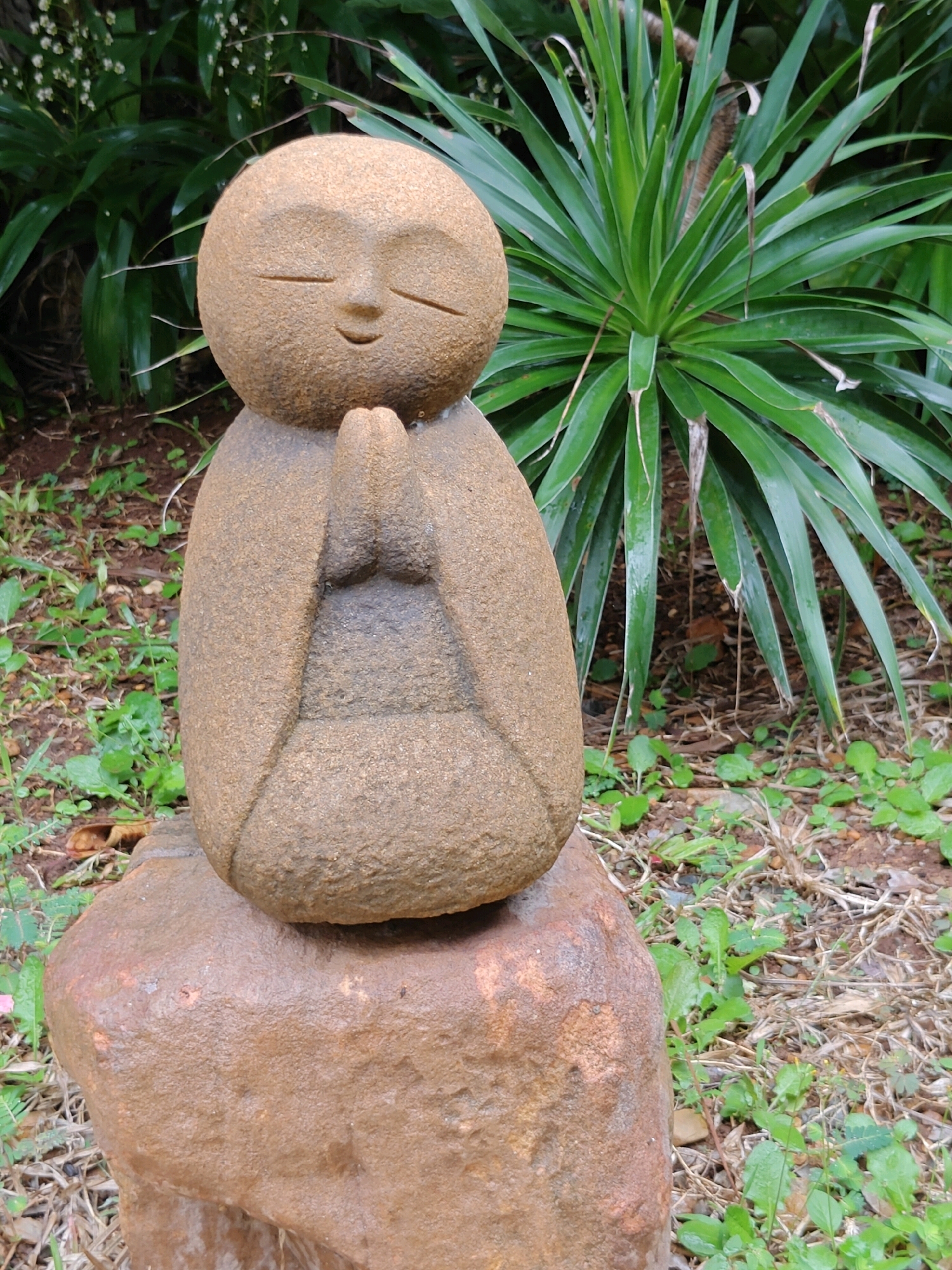 An upright, stone statue of a Jizo Bodhisattva is positioned in a garden setting, located in Mu Si, Thailand. The statue, carved from a light brown, porous stone, depicts a rounded figure with closed eyes, a subtle smile, and hands clasped in prayer. It rests upon a larger, rougher, reddish-brown rock base. The background consists of dense green foliage, including tall, spiky leaves of a plant behind the statue, and smaller green plants and dry grass on the ground. The lighting suggests an overcast day, as there are no harsh shadows.