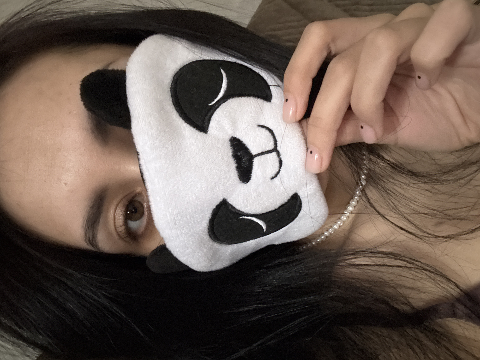 An eye-level close-up shot captures a person, likely female, lying down indoors while holding a white, plush panda-themed eye mask to their face. The person's right eye, with a brown iris and dark lashes, is visible, looking directly at the viewer. Their dark hair frames their face and spreads across the soft, light brown background, which appears to be a pillow or blanket. A delicate pearl necklace is visible around their neck. The person's right hand holds the eye mask, revealing light nude-colored fingernails, each adorned with a small black dot design. The panda mask features black embroidered eyes, nose, and mouth, giving it a cheerful expression. The lighting is soft and ambient, suggesting an indoor setting. The overall impression is one of relaxation or preparing for rest. No text is visible in the image. While the location context provided is Votkinsk, Russia, no specific visual cues confirm this.