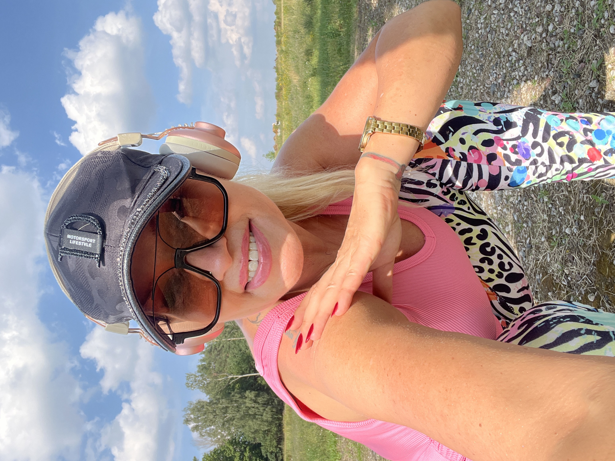 In Hodyszewo, Poland, a woman with blonde hair and red painted fingernails smiles at the camera. She is wearing a pink tank top, a black baseball cap with a diamond-studded brim, oversized sunglasses, pink headphones, and brightly patterned leggings. The woman is crouching in an outdoor setting with a field of grass and trees in the background under a blue sky with white clouds. She is touching her left shoulder with her right hand. A gold watch is visible on her left wrist.