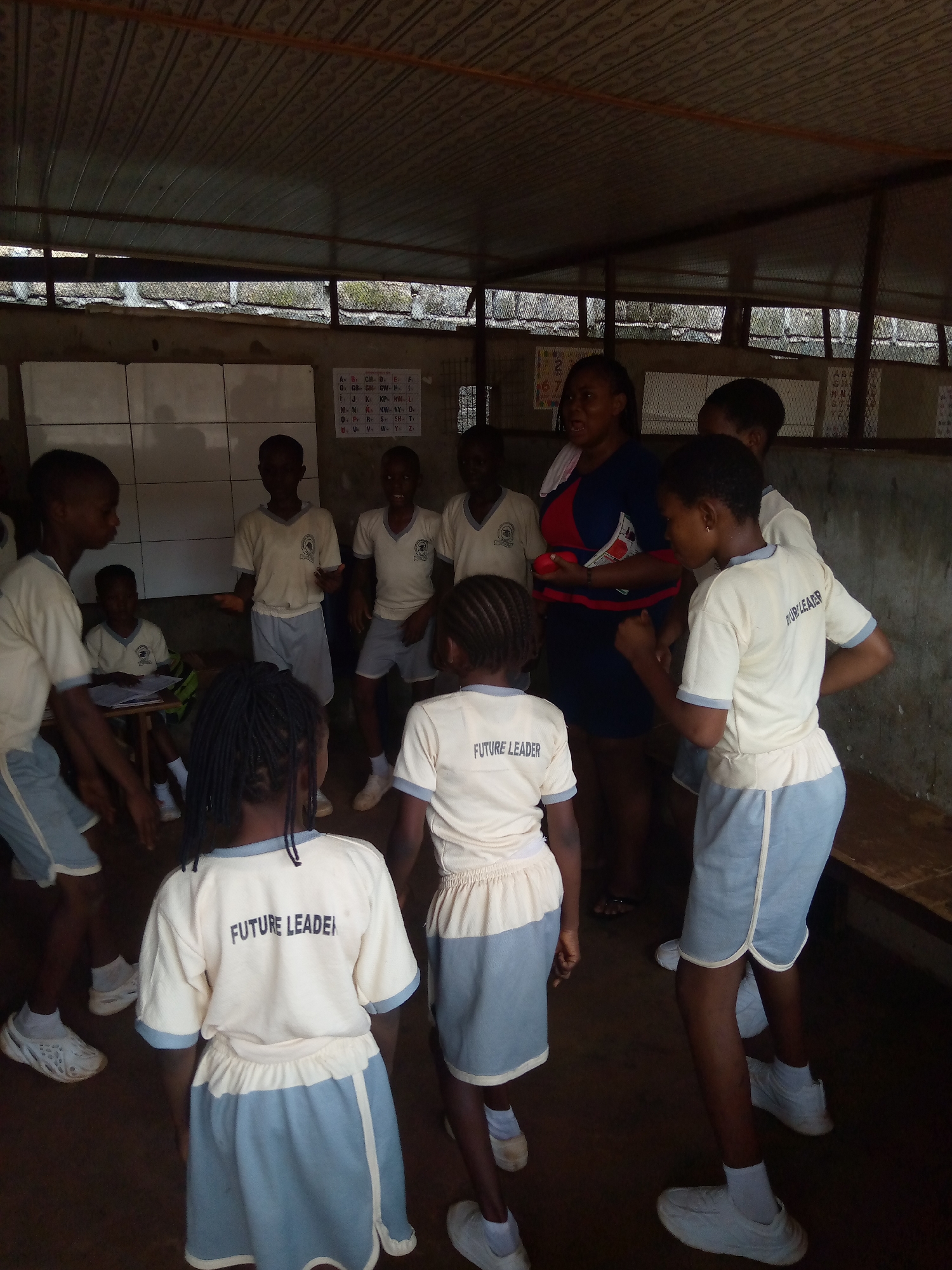 The image depicts an indoor scene, possibly a classroom or community hall, with a corrugated metal roof and concrete walls. Seven young students, dressed in identical light yellow t-shirts with "FUTURE LEADER" printed on the back and light blue shorts or skirts, are the central figures. They are either standing or sitting, engaged in an activity. A woman, likely an instructor or caregiver, stands in the background, wearing a blue and red patterned dress. The room appears somewhat rudimentary, with posters or charts on the walls, one of which displays letters of the alphabet. The lighting is dim, suggesting it might be daytime with limited natural light. The overall impression is of an educational or community gathering for children. The location is specified as Ukwu-Abwa, Nigeria.
