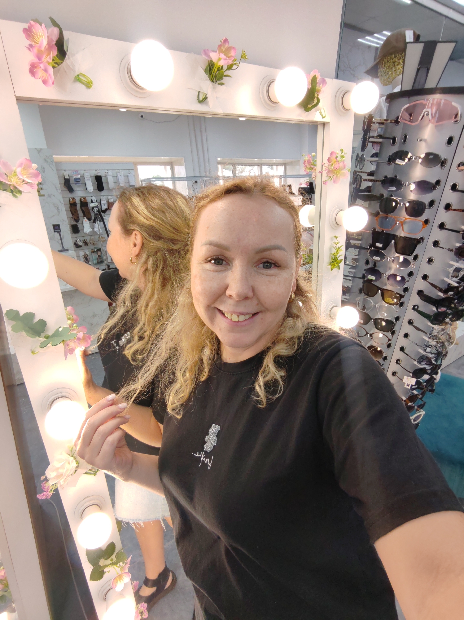 The image shows a woman taking a selfie in what appears to be a store. She is standing in front of a large mirror surrounded by light bulbs, a common feature of dressing room mirrors or makeup stations. The mirror reflects a display of sunglasses on a rack to her right and a rack of socks or hosiery to her left. She is wearing a black t-shirt and a denim skirt. She has blonde, curly hair and is smiling at the camera. In the background, through the mirror, another person can be seen. The lighting suggests an indoor environment, likely a retail space. The presence of sunglasses and hosiery indicates the store might sell clothing accessories or optical goods. The city of Chaikovsky, Russia is not explicitly indicated in the image, and there are no visual cues to confirm this location.