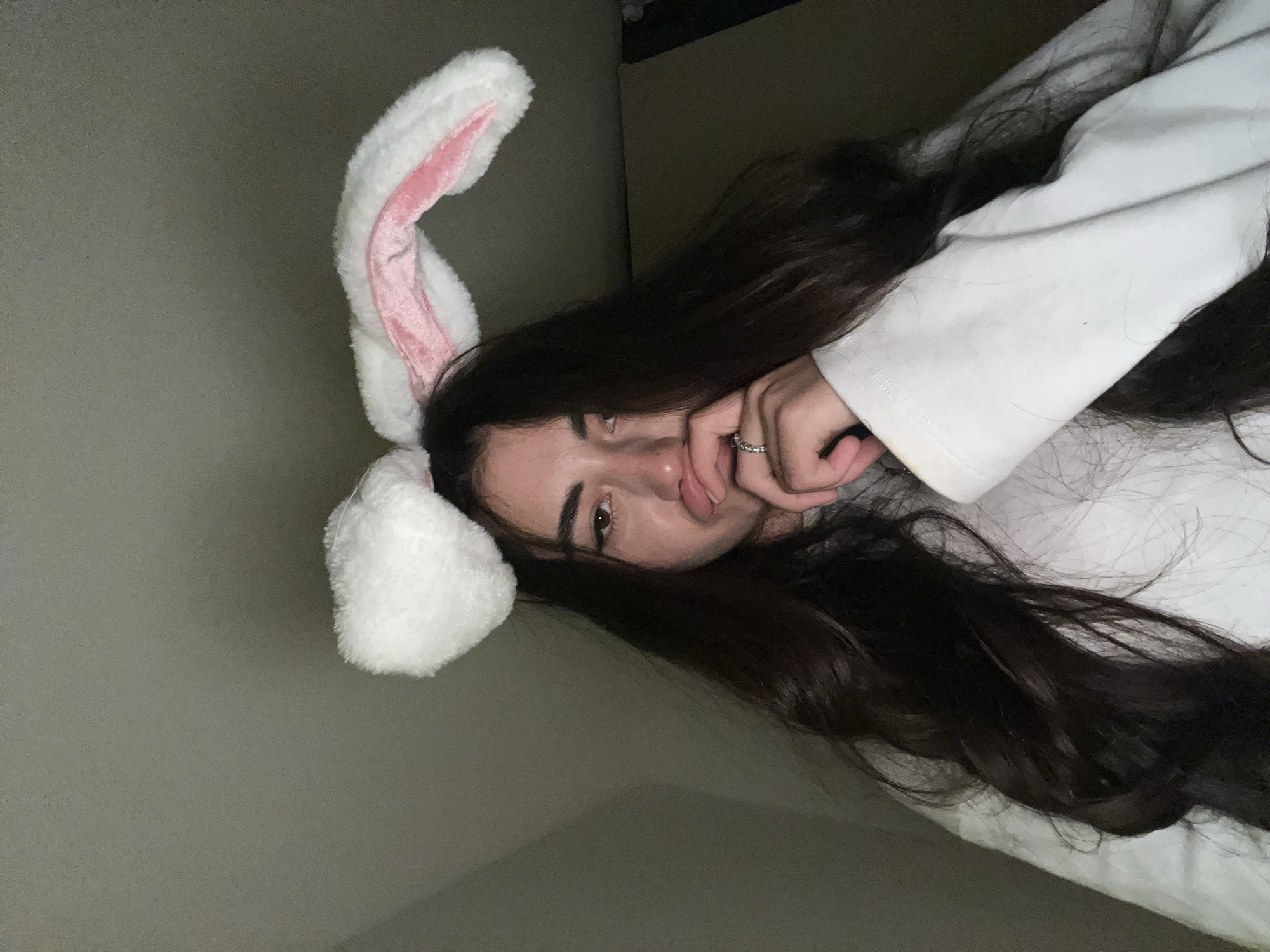 A young person with long, dark hair wears fluffy white and pink bunny ears. They are looking down and to the right, with their right hand touching their chin and mouth. They are wearing a white sweatshirt. The background is a dark, plain wall. The location is not specified in the image, and no information about Chotěšov, Czechia is present.