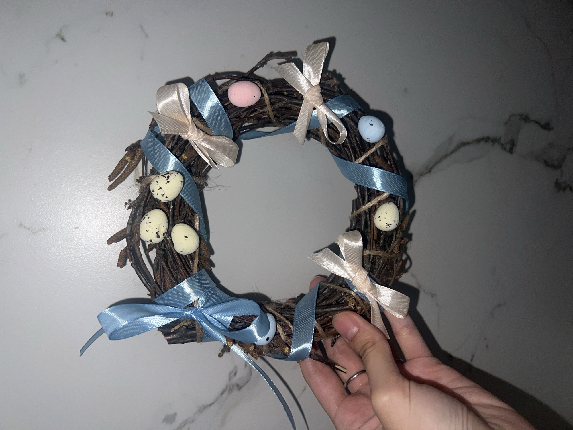 A close-up shot depicts a small, circular wreath resting on a surface that appears to be white marble or a material with a similar veined pattern. The wreath is constructed from interwoven twigs and adorned with pastel-colored miniature eggs (pink, blue, and speckled white) attached with twine. Light blue ribbon is woven throughout the wreath, accented by pale beige ribbon bows. A hand with a silver ring on one of the fingers is holding the wreath. No details indicate a location in Votkinsk, Russia. The environment seems to be indoors, possibly a home setting.