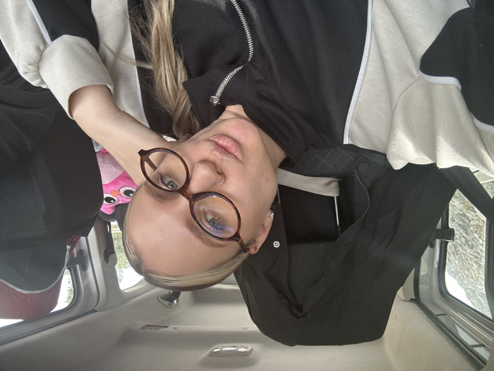 A fair-skinned individual is captured in a headshot selfie from inside a vehicle, located in Menteşe, Turkey. The subject has blonde hair, wears dark brown, oval-framed glasses, and a silver nose ring on their left nostril. They are dressed in a black and off-white/beige zippered jacket with white piping and are looking directly at the camera with puckered lips, their left hand resting casually near their head. The vehicle's interior features a light-colored ceiling and a black garment draped over the headrest directly behind the subject. Through the window to the subject's left, a rocky, possibly snowy, outdoor landscape is visible. To the subject's right, a pink owl-shaped plush toy is positioned on the adjacent seat, with green foliage visible through another window in the background.