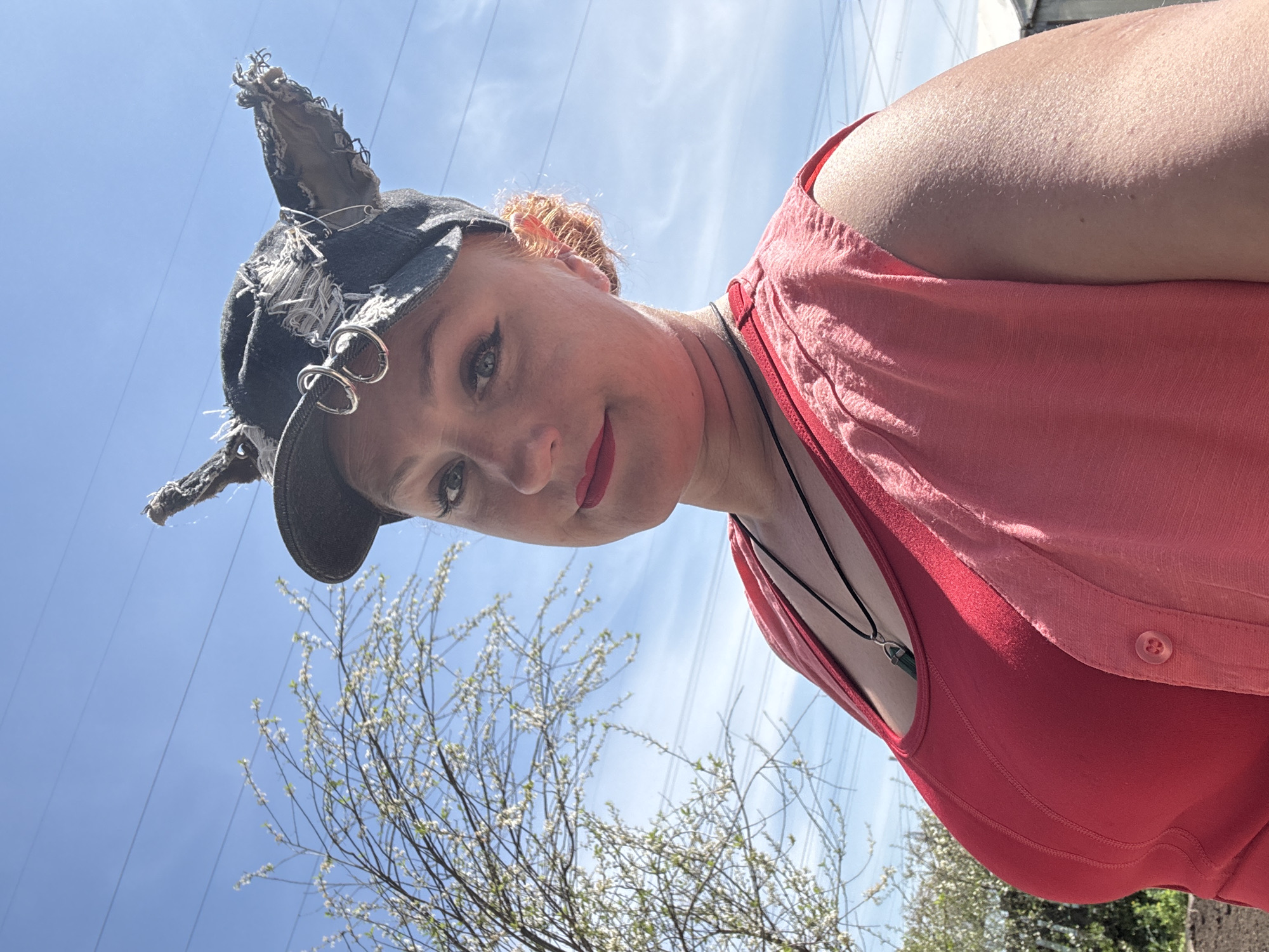 A woman with reddish hair is viewed from a low angle against a partly cloudy blue sky. She is wearing a red tank top under a lighter red button-up shirt. She has on a dark cap adorned with tattered fabric, metal rings, and a safety pin. A thin necklace with a green stone pendant is visible around her neck. The background includes bare tree branches with budding leaves and power lines. The location is potentially Chaikovsky, Russia.