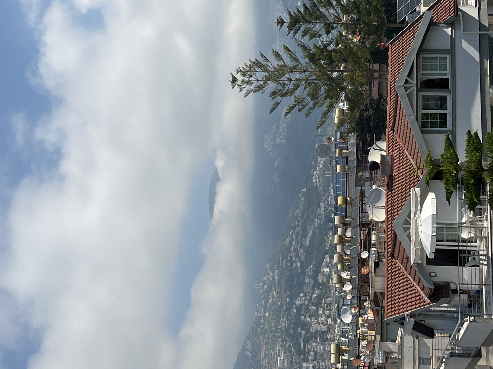 This image presents a wide panoramic view overlooking a mountainous coastal city, specifically Alanya, Turkey. The perspective is from an elevated position, looking down across rooftops and out towards distant mountains.
In the foreground on the right, a building with a distinctive red-tiled roof and white-framed windows is prominent. A large white patio umbrella is visible on a terrace of this building, along with some green trees, possibly conifers, that partially frame the scene.
Extending into the mid-ground, numerous rooftops of other buildings are visible, densely packed with an array of cylindrical solar water heaters and several white satellite dishes, suggesting a strong reliance on solar energy in the region. The urban area stretches across the hillsides, with countless houses and structures dotting the landscape.
The background is dominated by a range of mountains, partially veiled by soft, white clouds, creating a sense of depth and atmospheric haze. The sky above is bright, featuring a blend of clear blue patches and fluffy white clouds, indicating a daytime scene, likely under pleasant weather conditions.
No specific activities or people are discernible in the image; it captures a static, expansive landscape view. The abundance of solar water heaters and the architectural style are notable details that align with the provided location context of Alanya, Turkey. No visible text is present in the image.