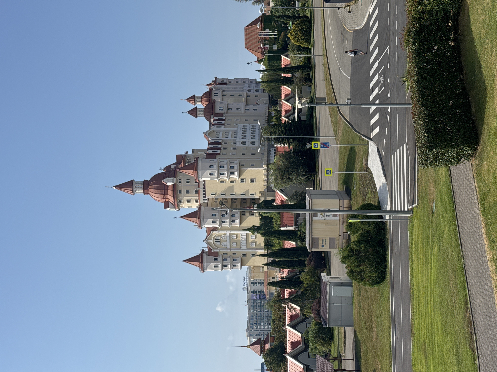 This is a photograph of a large, cream-colored, multi-story building with red-tiled, turreted roofs, resembling a castle, located in Sirius, Russia. In front of the castle are several smaller buildings with red-tiled roofs and an area with trees and greenery. A paved road with a pedestrian crosswalk is visible in the foreground, with a single person walking across it. Traffic signs and streetlights are also present. The sky is clear and blue. The overall scene suggests a modern resort or complex.