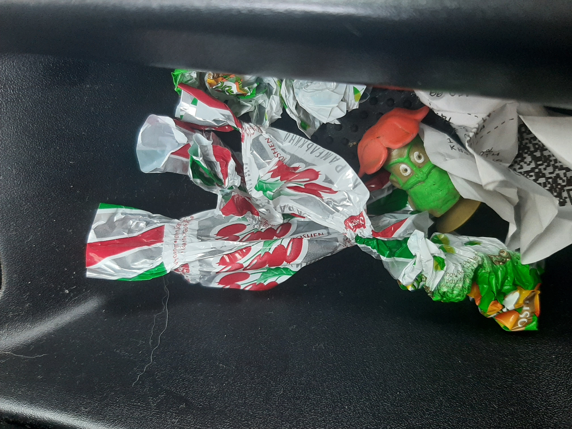 The image shows a close-up view of crumpled candy wrappers and a small toy figure inside a dark compartment, likely a car's glove box or center console. The wrappers are predominantly silver with red and green accents, featuring an image of cherries. Some text is visible on the wrappers, including "ROSHE" and what appears to be Cyrillic script.
The toy figure is green with a red, mushroom-shaped head and large, round eyes. It is positioned amongst the wrappers. A crumpled piece of white paper, possibly a receipt, is also visible in the background. The compartment's surface is textured and dark. The lighting suggests it is daytime, but no external environmental cues are visible. The overall impression is one of clutter and discarded items.