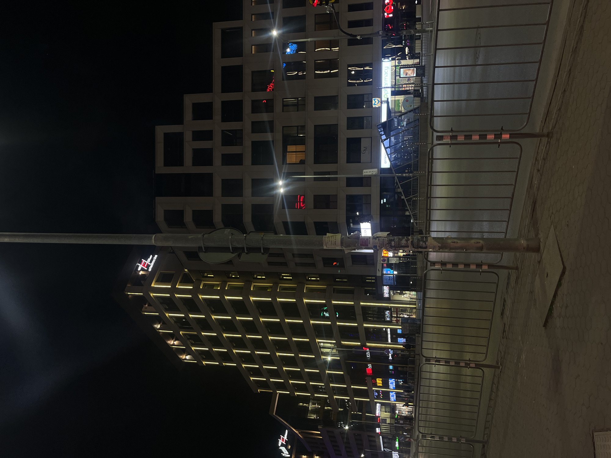 The image depicts a night scene in Sofia, Bulgaria. Two prominent buildings are visible. One is a modern building with a grid-like facade illuminated with yellow lights, topped with a logo that says "Grand Hotel Millennium". The other is a building with a boxy, stepped design, also lit up. Between the viewer and the buildings, there is a series of metal crowd control barriers along the sidewalk. The street is dimly lit. There is a light post with signs obscured by the camera angle.