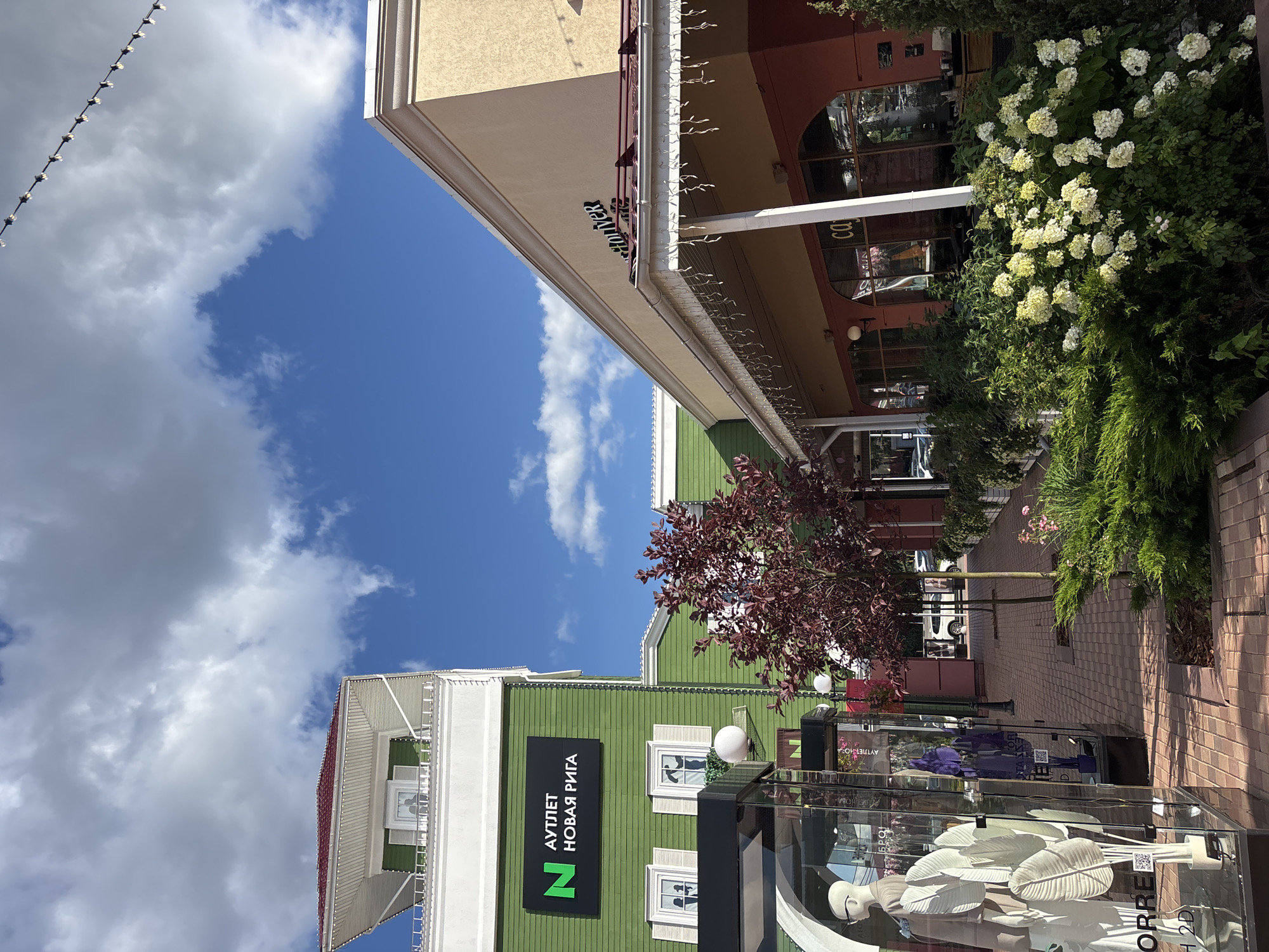 The image depicts an outdoor shopping area with two adjacent buildings under a bright, cloudy sky. The building on the left is painted green with white trim and features a prominent sign in Russian reading "N АУТЛЕТ НОВАЯ РИГА" (N OUTLET NEW RIGA). It has white-shuttered windows. In front of this building, a glass display window exhibits mannequins dressed in white clothing. A red-leafed tree stands between the buildings, and a brick pathway runs in front of them, lined with bushes and flowers. The building on the right is a lighter color, possibly beige or yellow, with a red awning and arched windows. Another sign, partially visible, reads "HOVER". Decorative string lights hang from the eaves of the building on the right. In the background, cars and other shoppers are visible, suggesting this is a public commercial space. The scene is well-lit, indicating daytime. This is the Outlet Village Novaya Riga, located in the Pokrovskoye settlement, Istrinsky District, Moscow Oblast, Russia, often referred to as part of the Moscow region, though Pokrovskoe itself is a settlement.