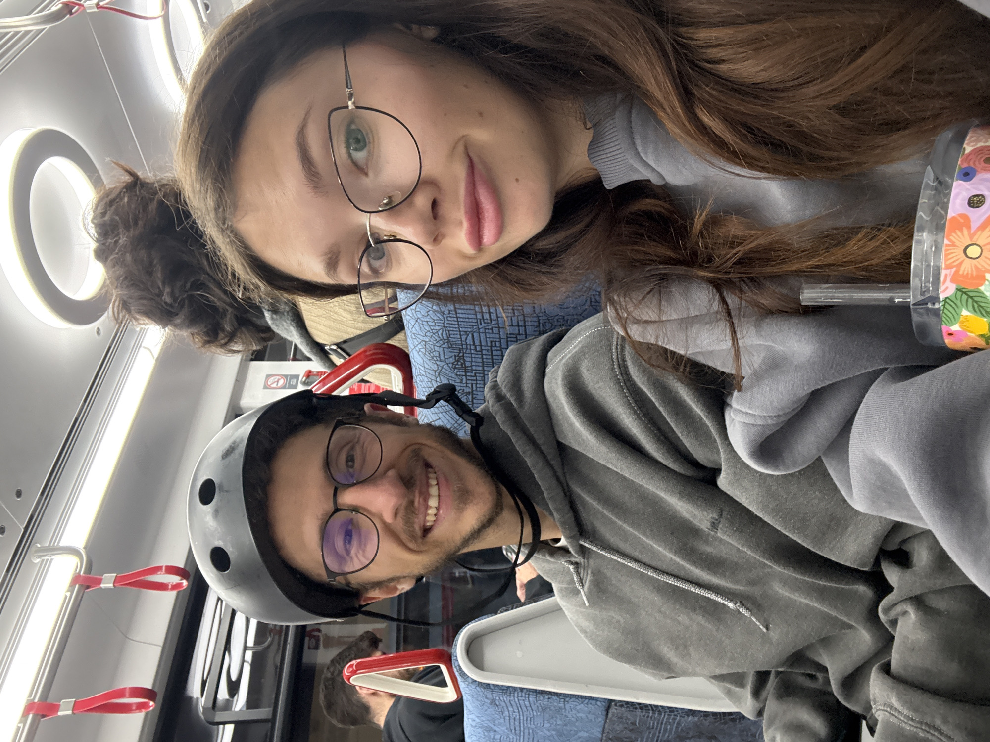 The image captures a selfie of two individuals, a woman and a man, inside a public transport vehicle, likely a train or tram in Tel-Aviv, Israel. The photo is oriented sideways, rotated 90 degrees counter-clockwise.
In the foreground, a woman with long, dark brown hair and large, fashionable glasses with thin black frames looks directly at the camera with a subtle smile. She has light-colored eyes, possibly green, and slightly parted pink lips. She is wearing a light grey or bluish-grey long-sleeved top. To her right, partially visible, is a clear tumbler with a straw, adorned with colorful floral patterns in orange, yellow, green, and white.
Beside and slightly behind the woman is a man, also smiling broadly at the camera, revealing his teeth. He wears a dark grey bicycle helmet with visible vents, round, dark-framed glasses, and a dark grey hooded sweatshirt. He has a beard and mustache.
The setting appears to be a modern public transport interior. Blue seats with a subtle geometric pattern are visible. The walls and ceiling are white or light-colored, featuring both circular and linear overhead lighting fixtures. Several red hanging straps or handles, typical for standing passengers, are prominently displayed in the background. Both subjects appear cheerful and relaxed, indicating they are enjoying their journey. The man's helmet suggests he might have been cycling. Faint, unreadable text or symbols can be barely discerned on a sign in the far background.