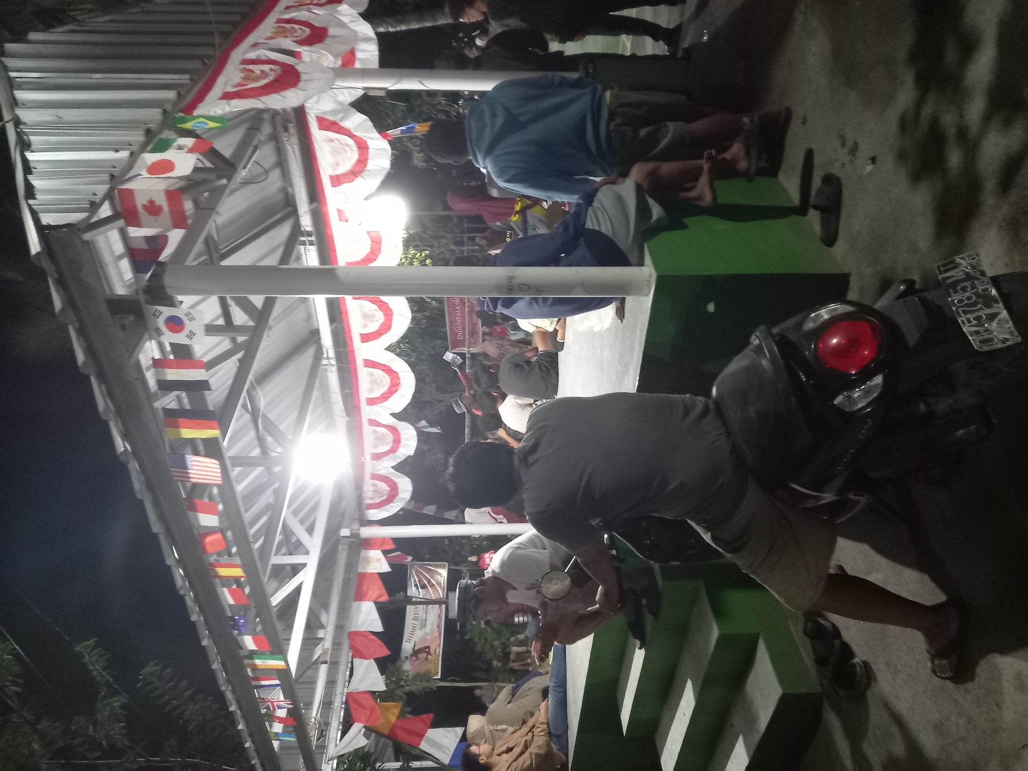 In Banyumulek, Indonesia, an outdoor gathering is taking place at night under a covered structure decorated with numerous national flags and red and white banners. Several individuals are present, some seated on green tiered steps and others standing. In the foreground, a person is sitting on a motorcycle, facing away from the camera. The motorcycle has a license plate that reads "DR 75785 MT". The environment appears to be an open area, possibly a community space or event venue, with lighting illuminating the scene.