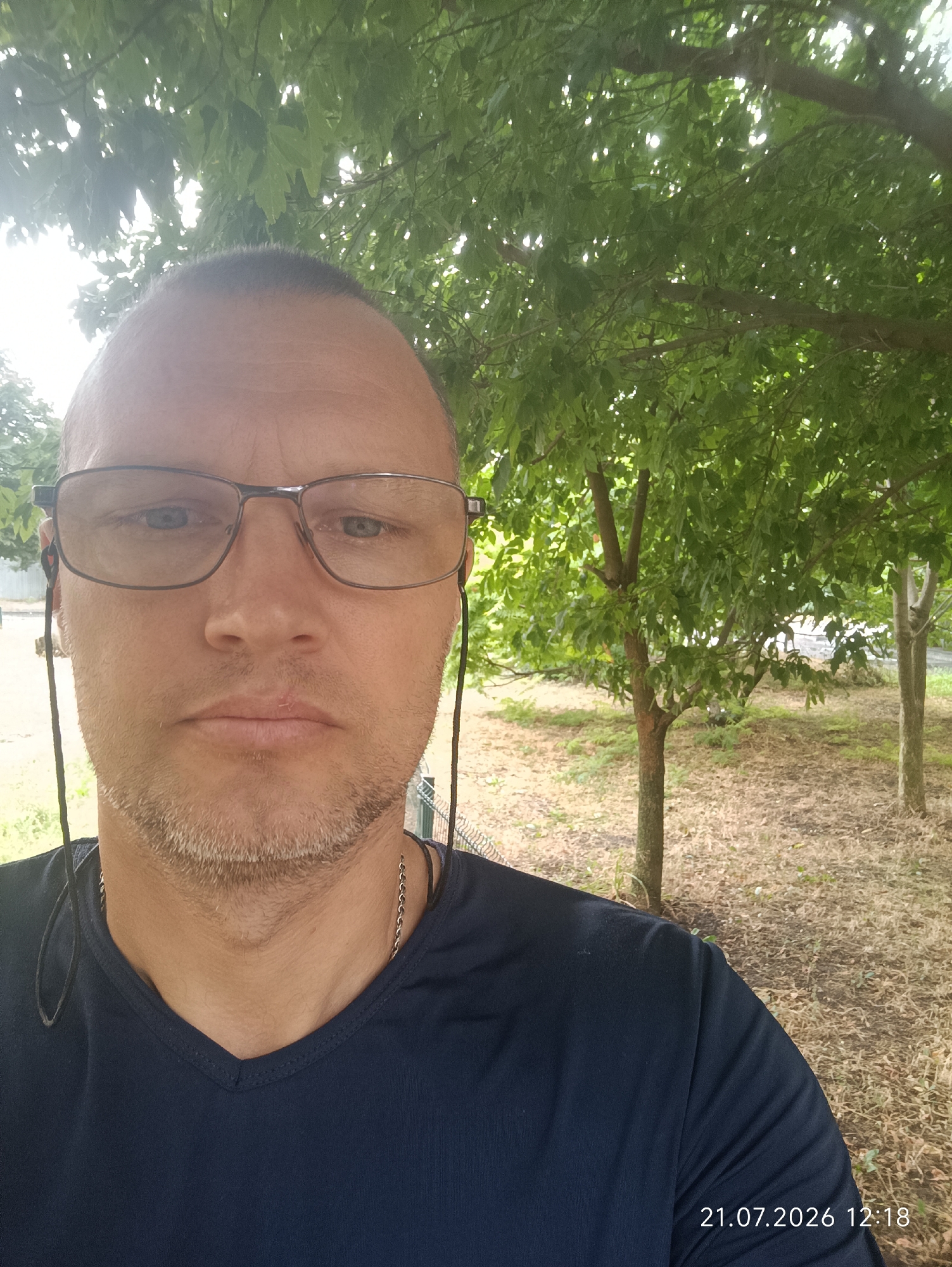 A man in his 40s with short hair and a light beard is taking a selfie outdoors. He is wearing glasses, a dark blue V-neck t-shirt, and earbuds. The man is looking directly at the camera with a neutral expression. The background consists of trees with green leaves, dry grass, and a fence. The image is timestamped "21.07.2026 12:18". The location is not specified.