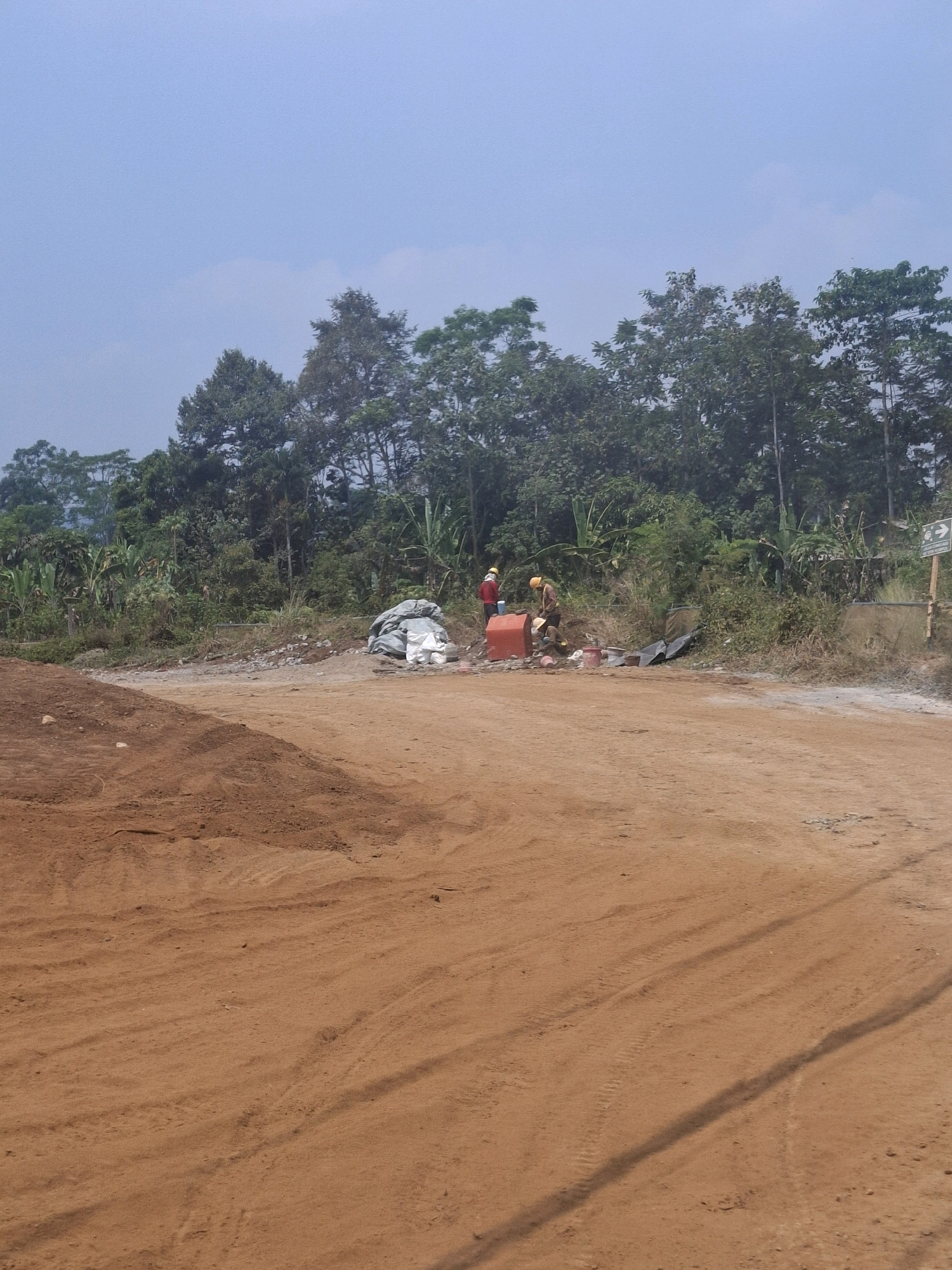 In Kutasirna, Indonesia, a construction site is depicted with an unpaved dirt road in the foreground, a pile of reddish-brown soil on the left, and a dense line of trees and foliage forming the background. Two individuals wearing hard hats and work clothes are present. One person, wearing a red shirt and a yellow hard hat, stands near a red barrel. The other person, in a yellow shirt and yellow hard hat, is engaged in some activity near the barrel and a pile of white bags. Several objects, including the red barrel, white bags, and scattered debris, are visible around the workers. A sign with an arrow pointing to the right is partially visible on the right side of the image. The sky is clear with some faint cloud formations. The overall scene suggests ongoing construction or land clearing activities.