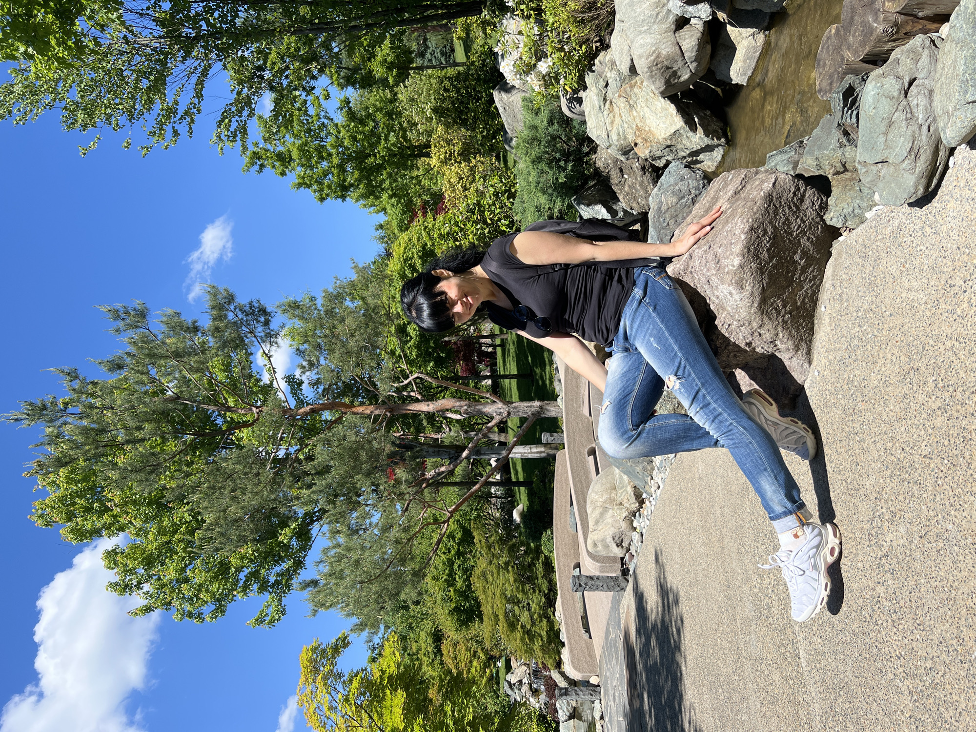 The image, correctly oriented, depicts a woman resting in a sunlit outdoor garden in Krasnodar, Russia. The primary subject is a woman with dark hair and bangs, wearing a black short-sleeved top, distressed blue jeans, and white athletic shoes with light brown accents. She is leaning or sitting on a large, light-colored rock positioned next to a narrow, shallow stream with murky green water. Her right hand rests on the rock, and she looks downward with a calm or contemplative expression. A black backpack is visible on her back, and sunglasses are tucked into her shirt collar.
The scene is a lush, vibrant park or garden, possibly designed in a Japanese style, featuring numerous large rocks, diverse green trees, and shrubs. The background is filled with rich foliage, showcasing various shades of green, with some trees also displaying hints of yellow. A wide, light-colored concrete or paved pathway runs through the foreground and beneath the woman, showing distinct shadows cast by her and nearby elements. The sky above is a clear, bright blue with a few scattered white clouds, indicating pleasant and sunny weather. The strong shadows suggest it is daytime, likely mid-morning or afternoon. No visible text is present in the image.