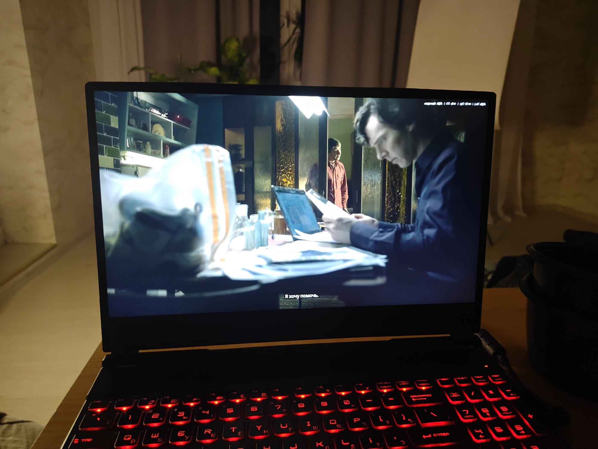 The image shows a laptop with a movie playing on its screen, displaying two men. One man, in the foreground, is wearing a dark blue shirt and is looking down at papers in front of him. Another man, in the background, is wearing a reddish-brown shirt and stands in a doorway. The laptop's keyboard has red backlighting. The scene is set indoors, with some shelving units visible on the left side of the screen, containing various items. The overall environment appears to be a dimly lit room. There is no visible information in the image to suggest the location is Krasnodar, Russia.