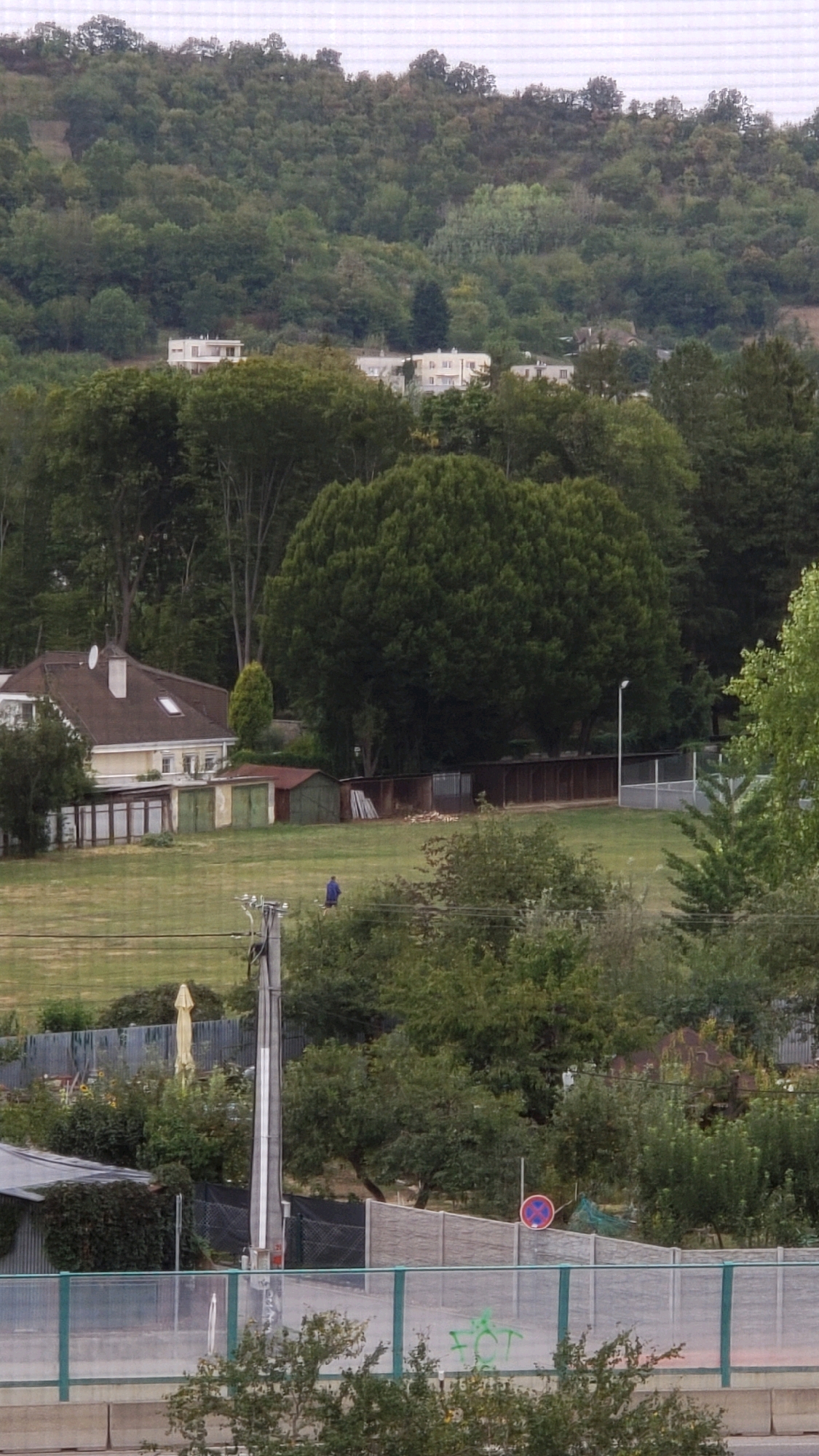 The image depicts a residential area with a hilly, wooded backdrop. In the foreground, a paved area with a green railing is visible, with graffiti on the railing. Beyond this, a large grassy field stretches out, containing a lone individual wearing a blue shirt walking across it. To the left of the field is a light-colored house with a dark roof, accompanied by several small outbuildings, possibly garages. Further into the background, more houses and apartment buildings are nestled amongst the trees on the hillside. A streetlamp is visible in the middle ground. The overall impression is a suburban or semi-rural landscape.