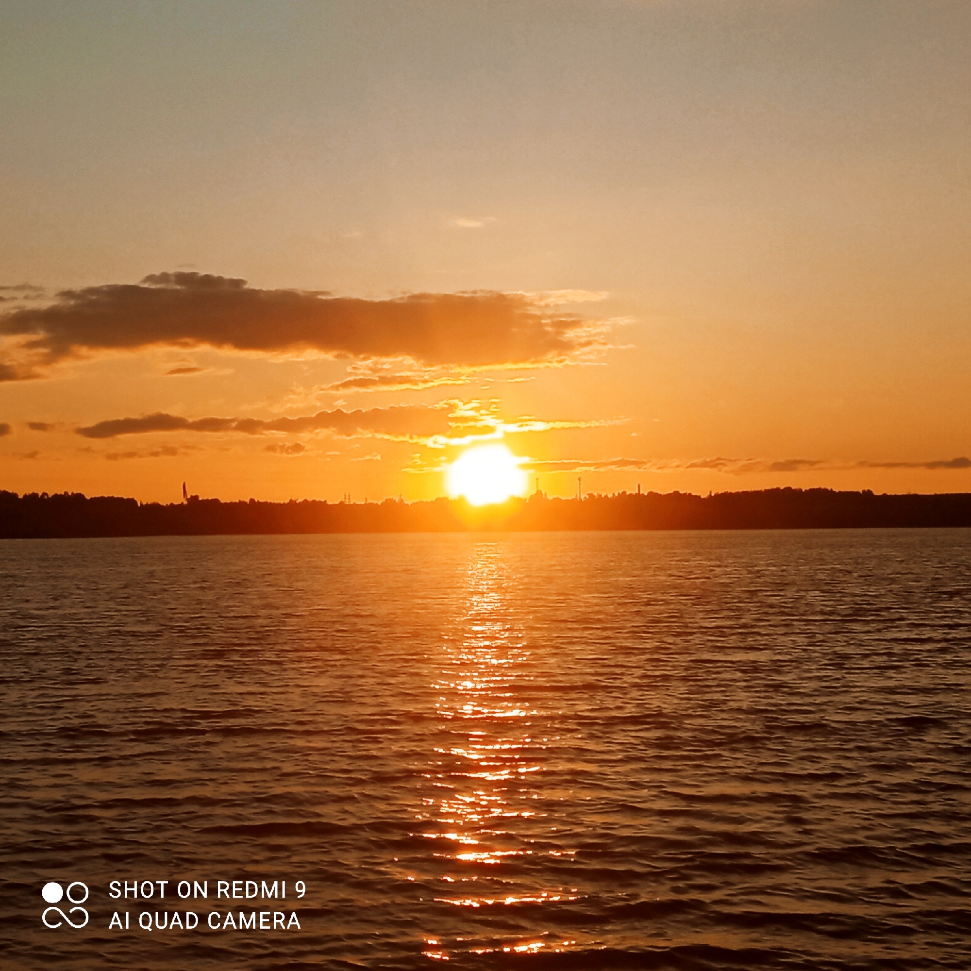 The image shows a sunset over the Kama River near Chaikovsky, Russia. The sun is low on the horizon, casting a warm, orange glow across the sky and water. Clouds are scattered across the sky, some tinged with pink and orange. A silhouetted treeline is visible along the far bank of the river, with some structures possibly indicating industrial or urban presence. The water is rippled, reflecting the vibrant colors of the sunset. In the lower left corner, a watermark states "SHOT ON REDMI 9 AI QUAD CAMERA".