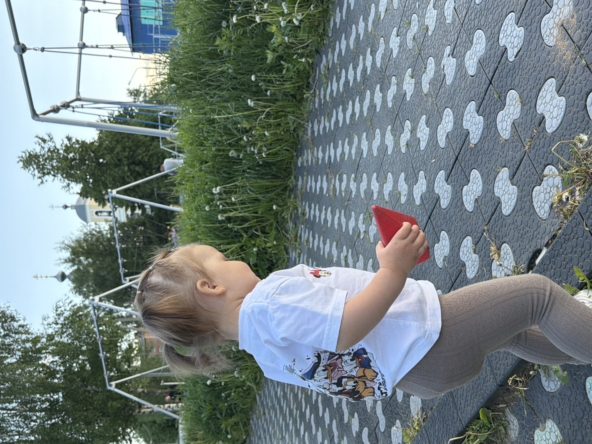 A young child stands on a paved surface at a playground in Sin'kovo, Russia. The child is wearing a white t-shirt with cartoon characters and light brown leggings, holding a red object in their hands. The ground is paved with black tiles with a white clover-shaped pattern. To the left is a grassy area with dandelions and a metal playground structure, partially visible. In the background, trees and the dome of a church can be seen under a bright sky.