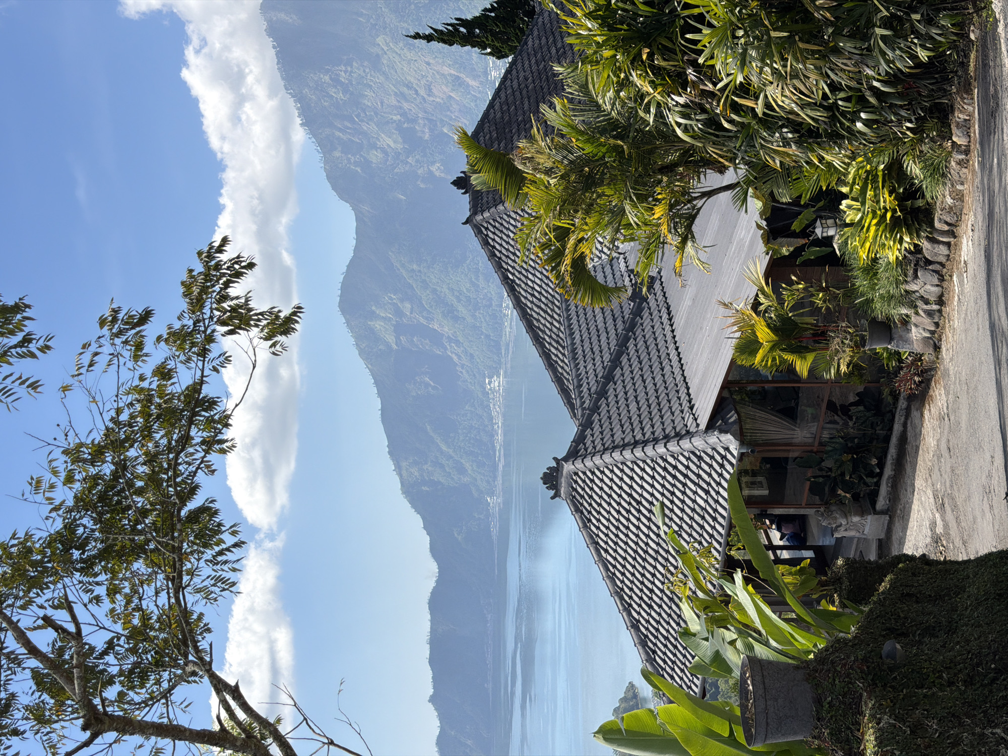 The image captures a serene view from a vantage point in Kintamani, Indonesia. In the foreground, lush green foliage and tropical plants surround a grey tiled roof of a building, likely part of a restaurant or guesthouse, with traditional Balinese architectural elements. A pathway leads towards the building, flanked by more vegetation and a stone wall. In the middle ground, the expansive, tranquil waters of a lake are visible, reflecting the clear blue sky. Beyond the lake, a majestic mountain range dominates the background, its slopes covered in dense greenery and dotted with scattered settlements. A thin plume of smoke rises from the lake, hinting at distant activity. The scene is bathed in bright sunlight, suggesting a pleasant day. A few people are seated inside the building, their presence subtly noted.