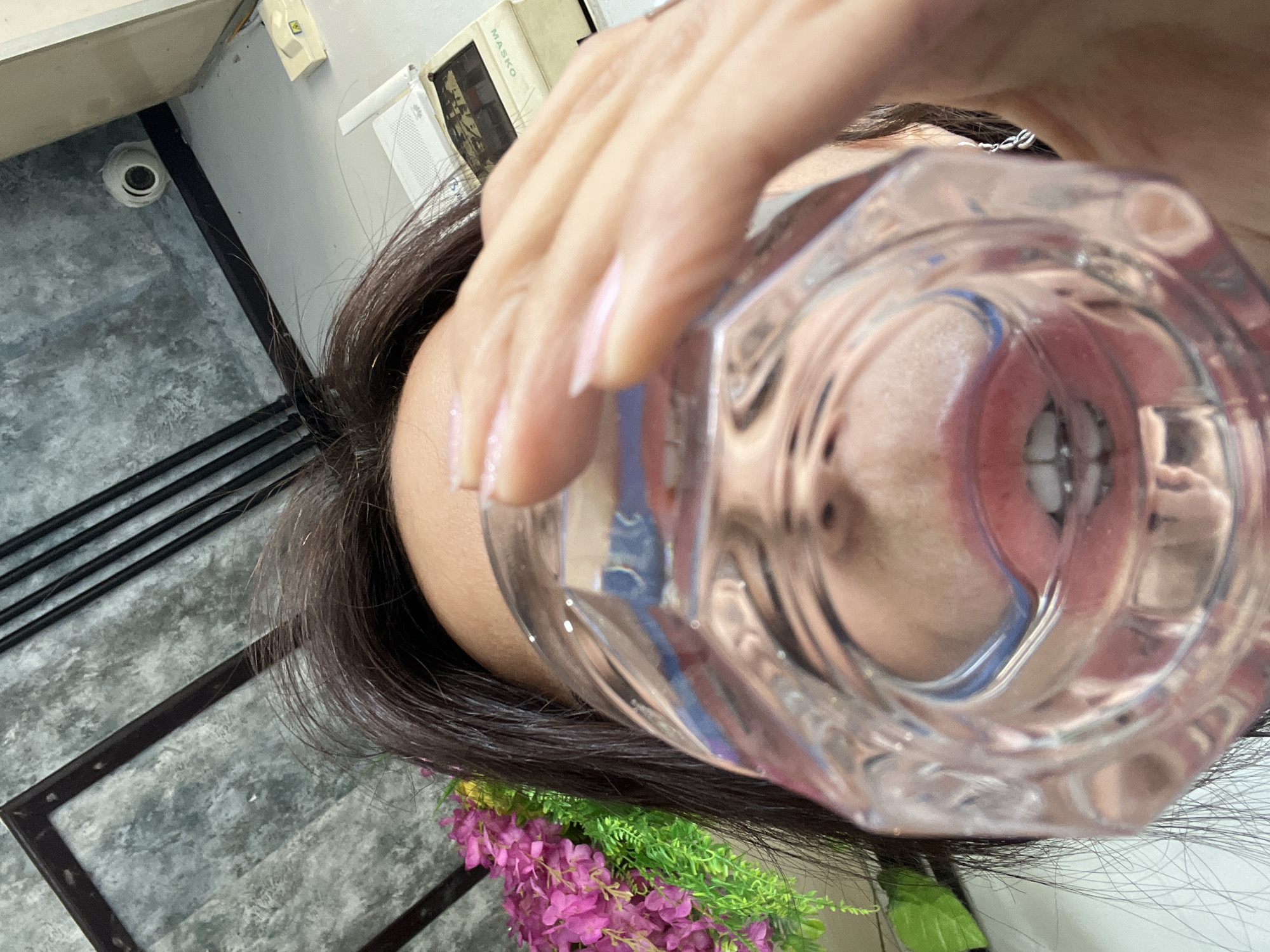 A woman holds a drinking glass filled with liquid in front of her face. The glass distorts her face, showing her teeth and facial features in a warped perspective. The woman has dark hair. Part of a floral arrangement with pink and green elements is visible near her hair. In the background, the flooring appears to be patterned gray and black. The presence of Indonesian books on the back indicates that the image was taken in Kuta Selatan, Indonesia. The location is a domestic interior setting, judging by the flooring and interior design. The subjects are primarily focused on the woman and the glass of liquid.