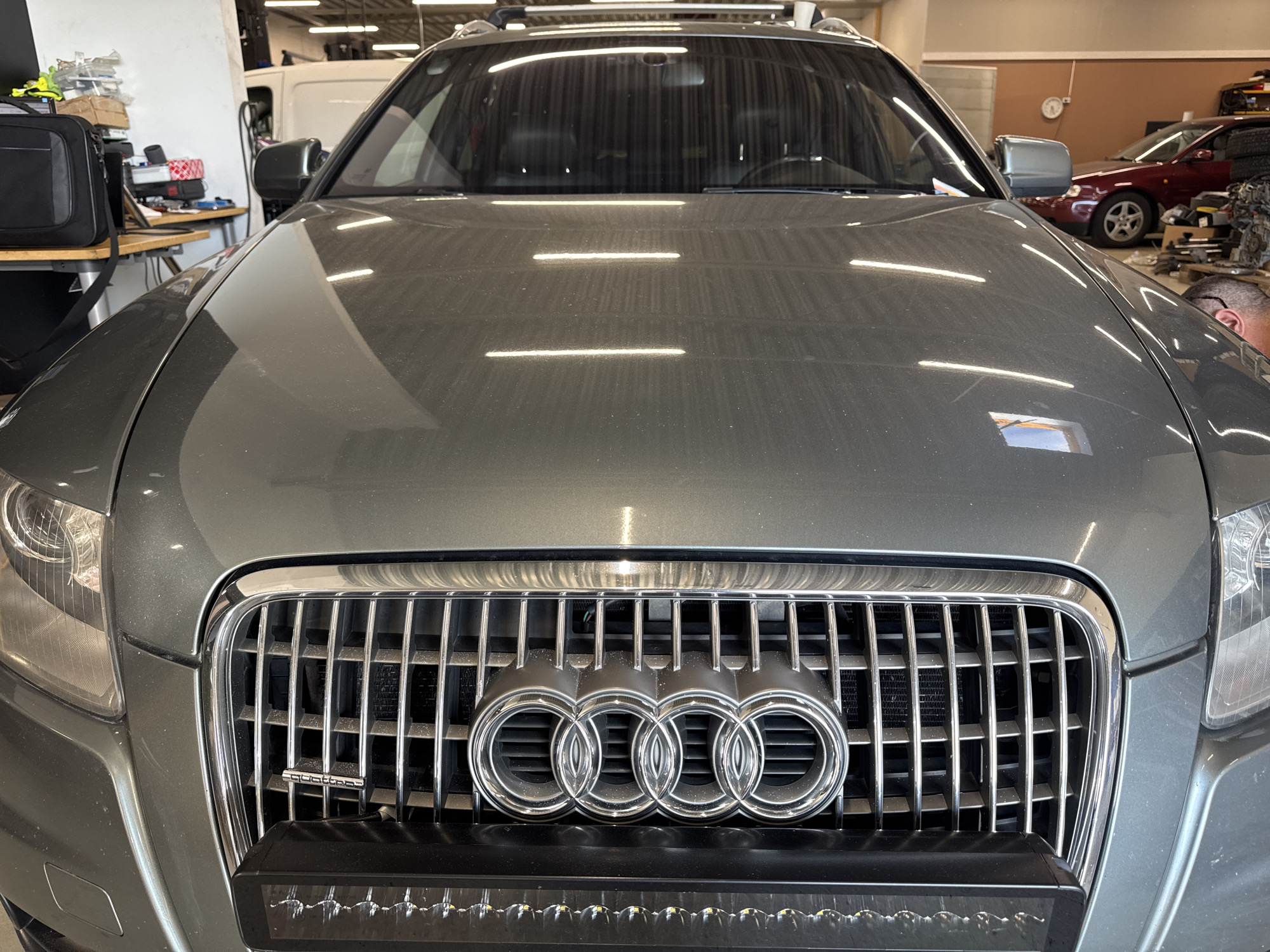 The image shows the front of a grey Audi SUV parked indoors, likely in a workshop or garage. The vehicle has a prominent chrome grille with the Audi rings logo and a "quattro" badge. A long LED light bar is mounted below the grille. The background shows various items, including a desk with bags and boxes, a white van, and another car. The overall impression is of a well-lit indoor space, possibly a car repair shop. There is no clear indication of the location being Sandnes, Norway.