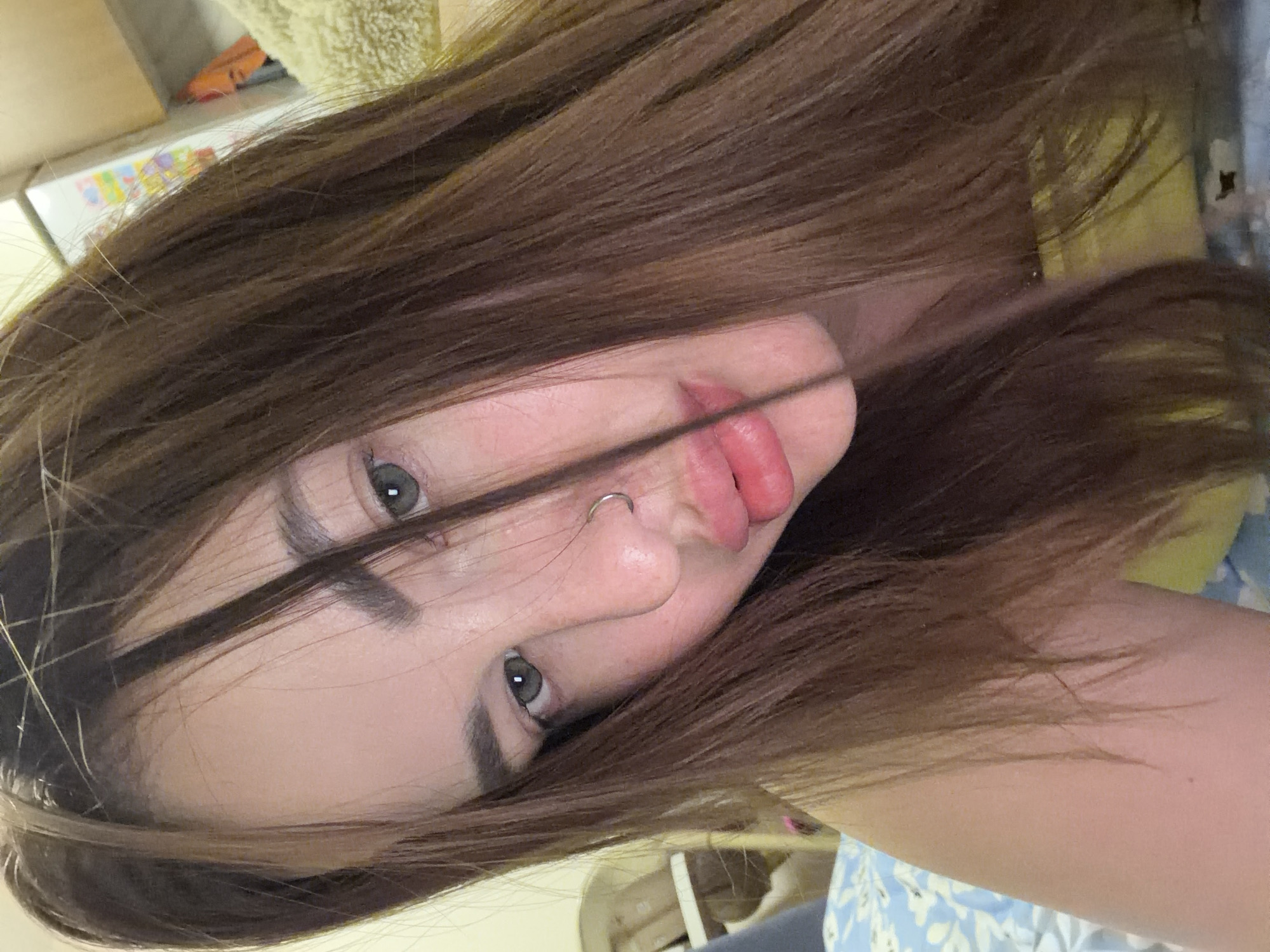 A close-up, tilted selfie of a person with long, straight brown hair partially covering their face. The person has light skin, grey eyes, a nose piercing, and slightly parted lips. The background is blurry and appears to be an indoor setting with shelves, a yellow object, and blue patterned fabric. There is no information to specify the location of the person. The information that the city of Krasnodar, Russia is irrelevant to the content of the image.