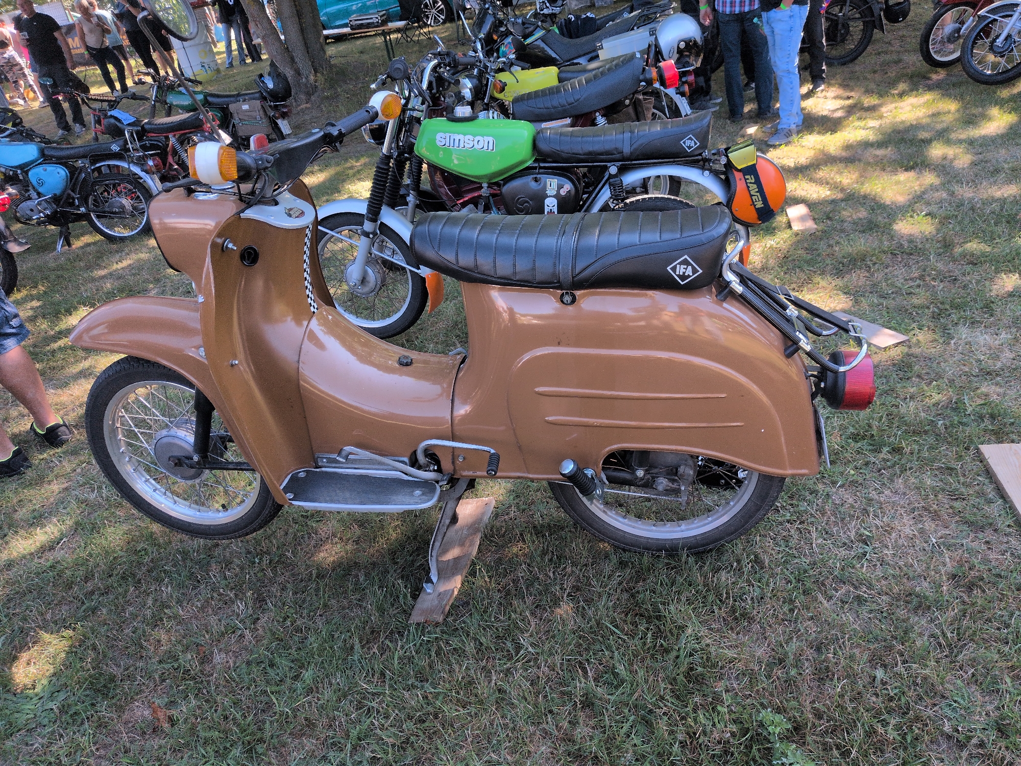 The image depicts an outdoor event, likely a motorcycle gathering, taking place on a grassy area with some trees in the background. The foreground is dominated by a brown scooter, possibly a Simson model, with its front wheel on the left and the rear wheel visible on the right. The scooter has a black seat with an "IFA" logo. Behind the brown scooter, other vintage motorcycles are parked, including a green Simson with the brand name prominently displayed on its fuel tank. Several people are visible in the background, some standing and others walking, dressed in casual clothing. The environment suggests a sunny day, with shadows cast on the grass. The location is identified as Hohenferchesar, Germany.