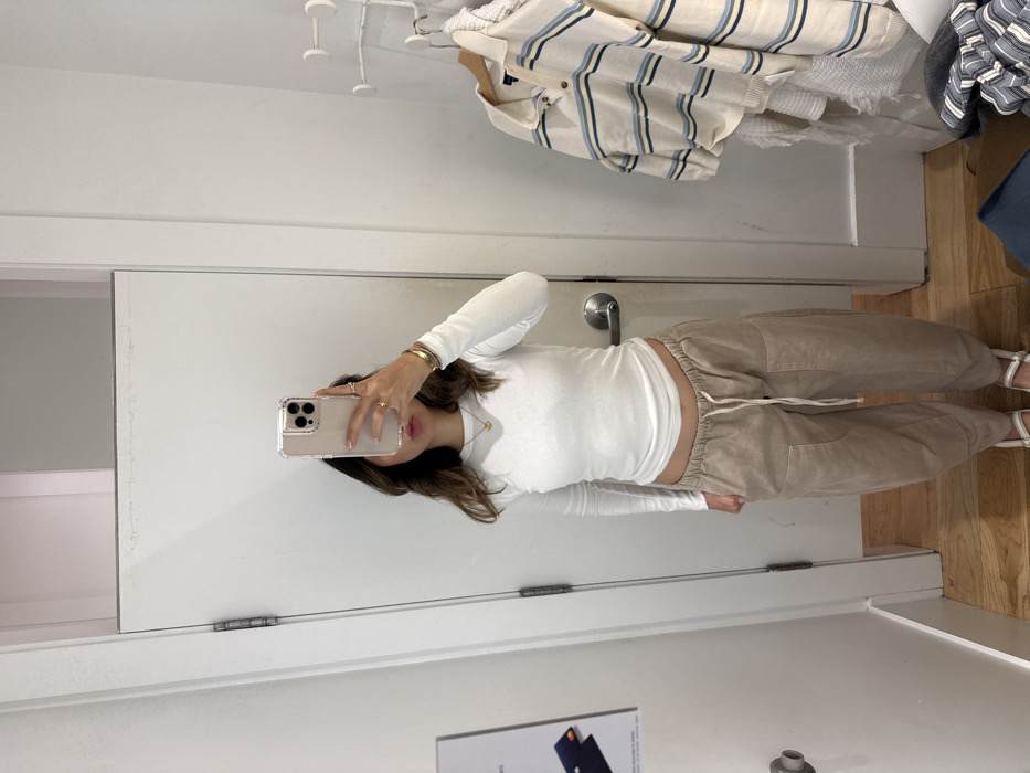 A woman takes a mirror selfie in a dressing room. She wears a white, long-sleeved shirt and beige drawstring pants with white sandals. Clothes hang on hooks above, including a striped polo and folded garments. A closed dressing room door and a partial view of an advertisement are also visible. The image is taken at an angle and appears slightly rotated counterclockwise.