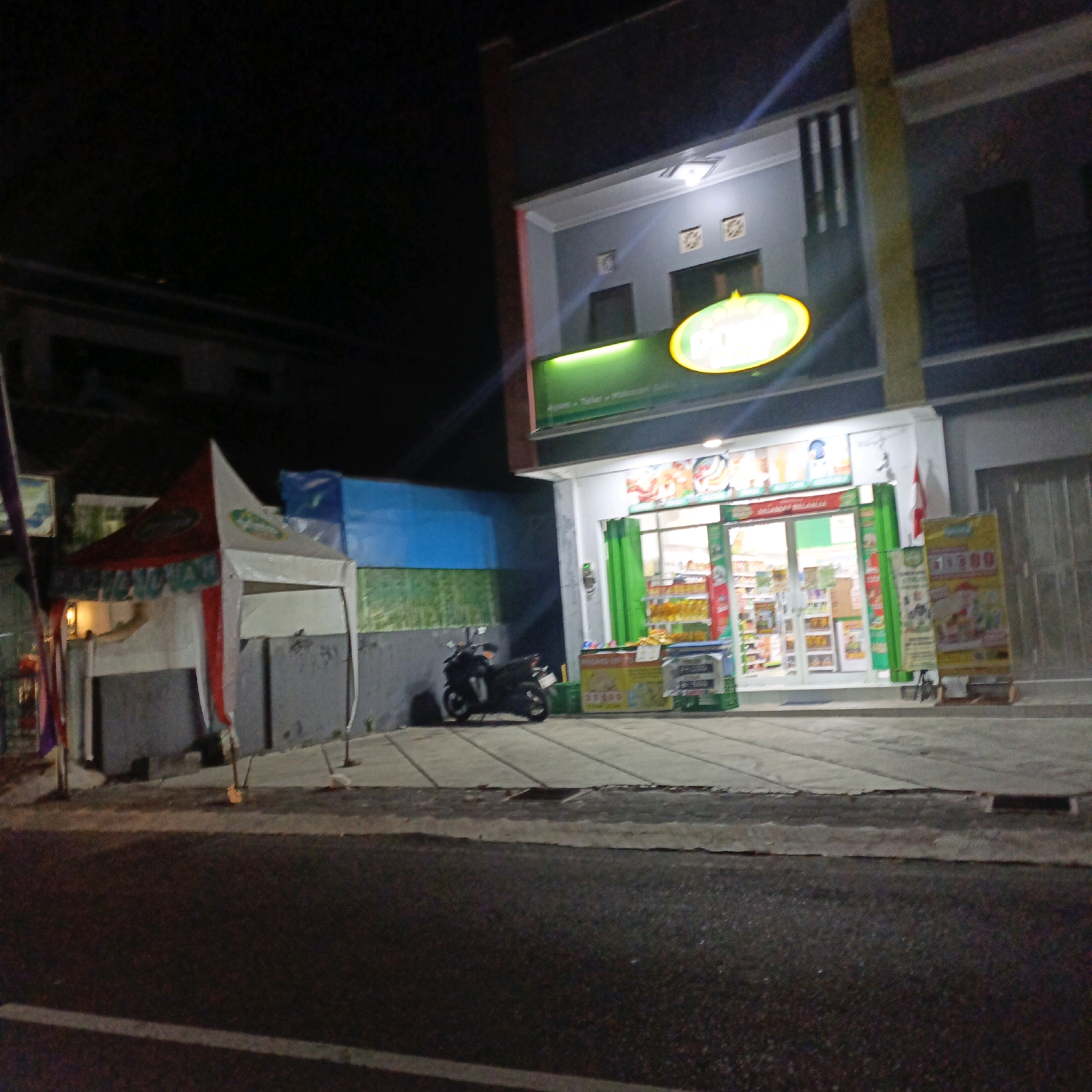 At night in Salatiga, Indonesia, a well-lit convenience store with a green and yellow oval logo is the central focus. The store has a wide glass storefront displaying shelves stocked with merchandise, and green curtains are visible inside. A motorcycle is parked on the sidewalk in front of the store, to the left. To the left of the store, a red and white tent is set up, with some banners and what appears to be a light source inside. The street in front of the store is asphalt, with a white line marking the edge. The area around the store appears to be a commercial or residential street with adjacent buildings. The overall lighting suggests it is after dark, with artificial lights from the store and surrounding areas illuminating the scene.