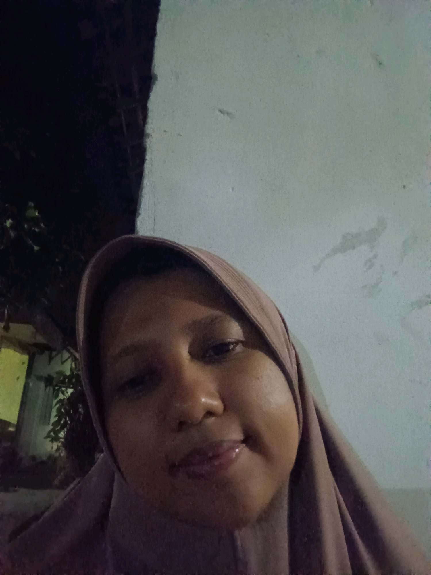 The image is a close-up selfie of a person wearing a hijab. The person's face is partially visible, with their eyes, nose, and mouth in focus. The background is divided into two distinct areas: the left side is dark and shows foliage and what appears to be a building structure with yellow lighting, while the right side is a lighter, textured wall. The lighting in the image is dim, suggesting it was taken at night or in a low-light environment. The composition is centered on the person's face. There is no discernible activity or interaction taking place. The location is identified as Canga'an, Indonesia.