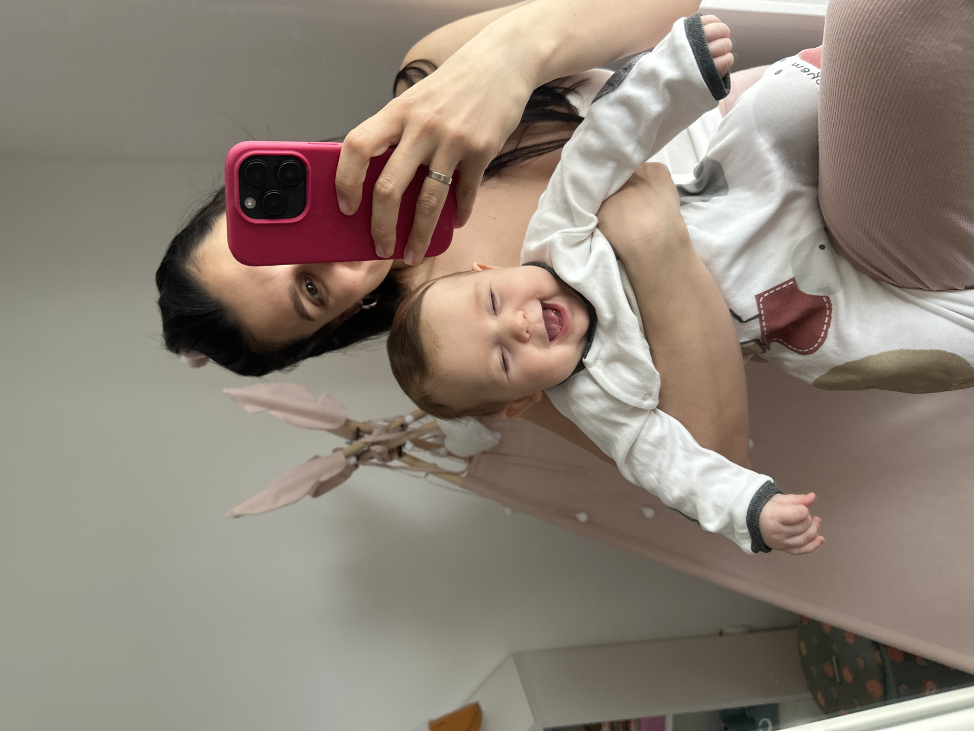 The image is a mirror selfie taken indoors, featuring a woman holding a baby in what appears to be a home in Chaikovsky, Russia. The woman, with dark hair and wearing a dark top and light pink ribbed pants, holds a bright pink smartphone with three camera lenses, visible in the upper part of the frame. A silver ring is visible on her left hand. She is holding an infant, who is wearing a white long-sleeved onesie with a cartoon animal design and dark grey trim on the cuffs. The baby is captured mid-laugh or joyful shout, with eyes squeezed shut and mouth wide open, conveying pure happiness. The woman's eye, visible in the reflection, shows a gentle expression.
In the background, a light pink play tent (teepee) with wooden poles is partially visible. Below it, a white piece of furniture, possibly a shelf or dresser, can be seen, along with a patterned storage bin with orange dots to the right. The lighting suggests daytime, creating a soft, warm ambiance for this tender and playful moment between mother and child. There is no visible text in the image.