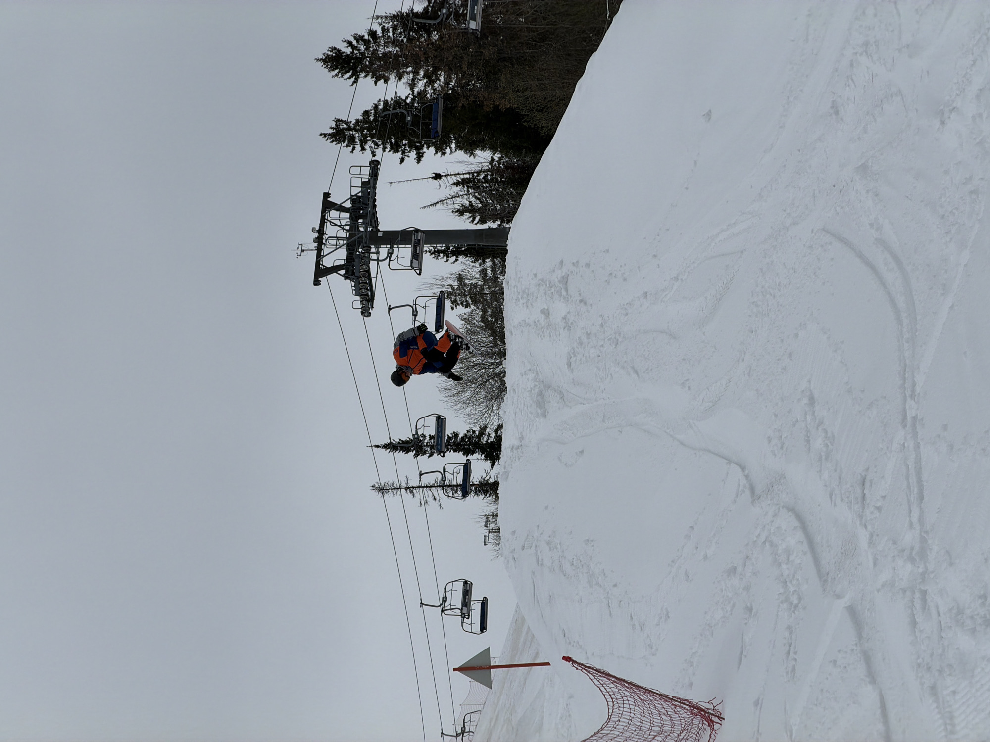 The image depicts a skier in mid-air, performing a trick, likely at the Nechkino ski resort in Russia, given the prompt. The skier is wearing orange and black clothing. A ski lift is visible in the background, composed of towers and chairs. The environment is snowy, with snow-covered ground in the foreground and trees in the background. The sky is overcast. A red safety net and a triangular marker are positioned on the slope.