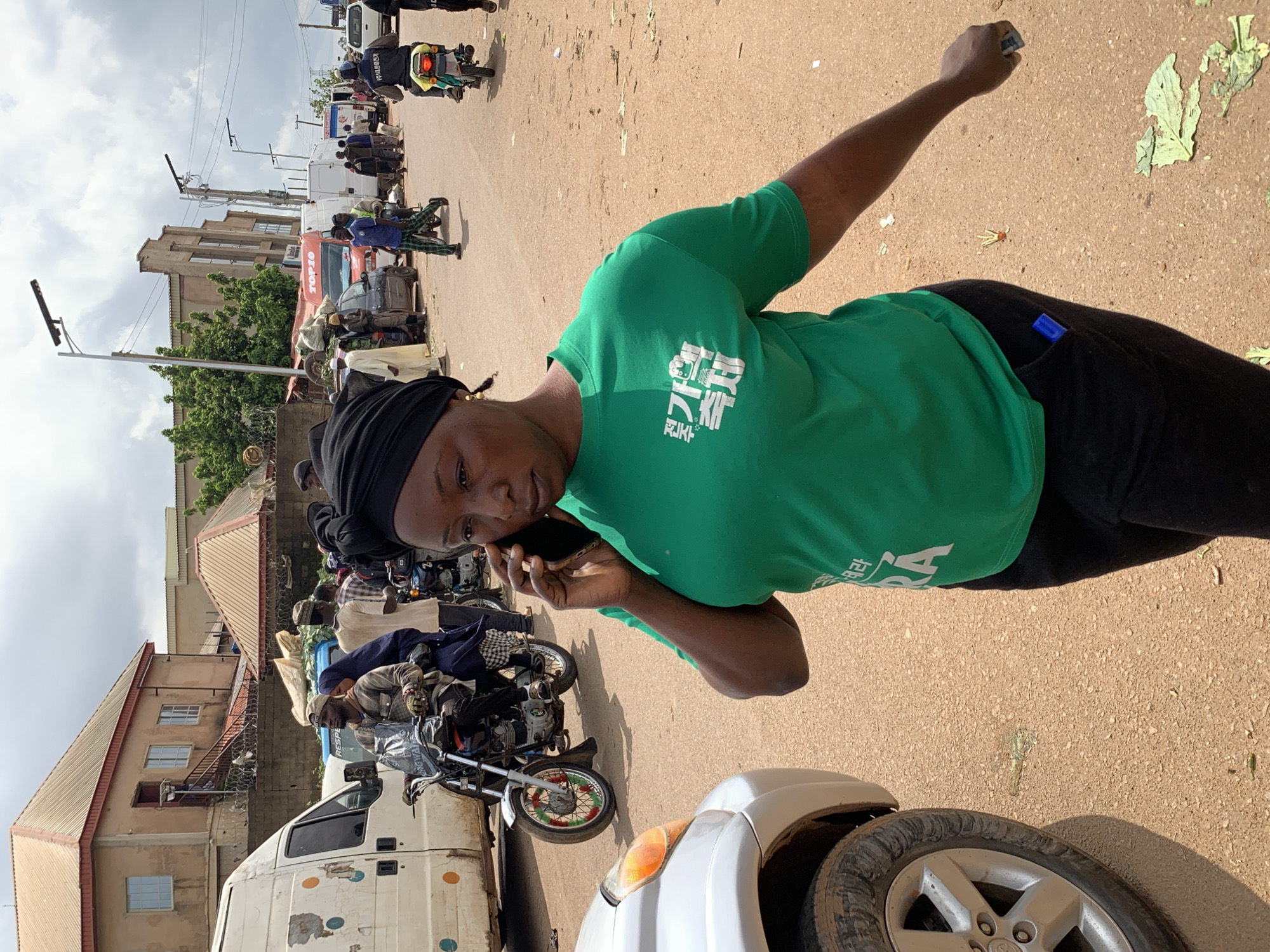 The image depicts an outdoor market scene in Gura Top, Nigeria. A woman in a green t-shirt and black headscarf is on the phone in the foreground, with a silver car to her left. Behind her, various people and vehicles are visible, including motorcycles, vans, and cars. Some individuals are on motorcycles, while others are walking or standing in the bustling market. Buildings line the background, suggesting an urban or semi-urban setting. The ground is a dirt road, typical of many Nigerian streets. Scattered debris, possibly organic matter, is present on the ground. The overall impression is one of a busy, active marketplace with a blend of traditional and modern elements.