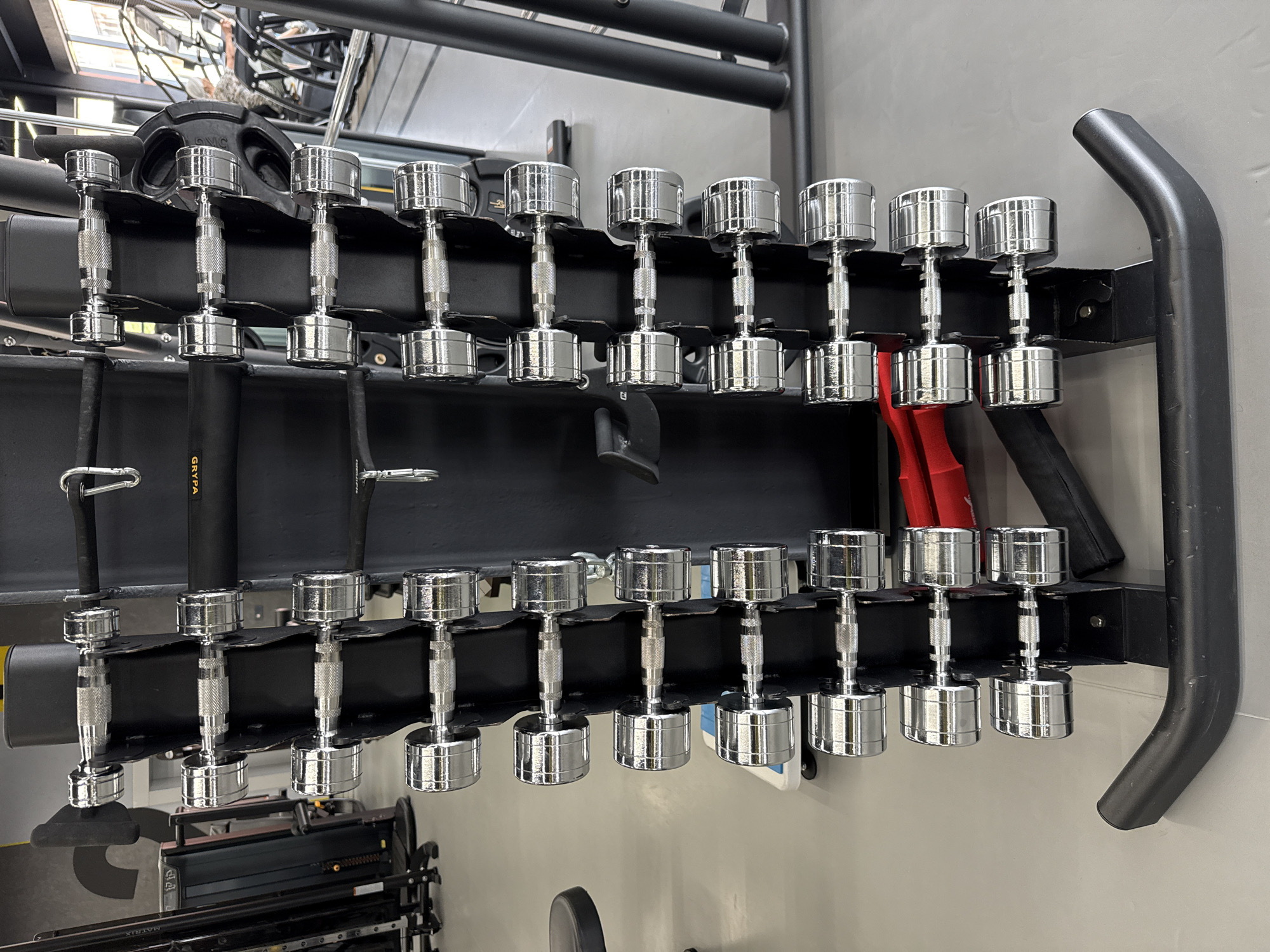 The image depicts a weight rack filled with chrome dumbbells. The dumbbells are arranged in two rows on a black metal rack. The rack is positioned against a light gray wall. In the background, other gym equipment is visible, including weight machines and bars. The setting appears to be a gym or fitness center. The image does not contain information about Votkinsk, Russia.