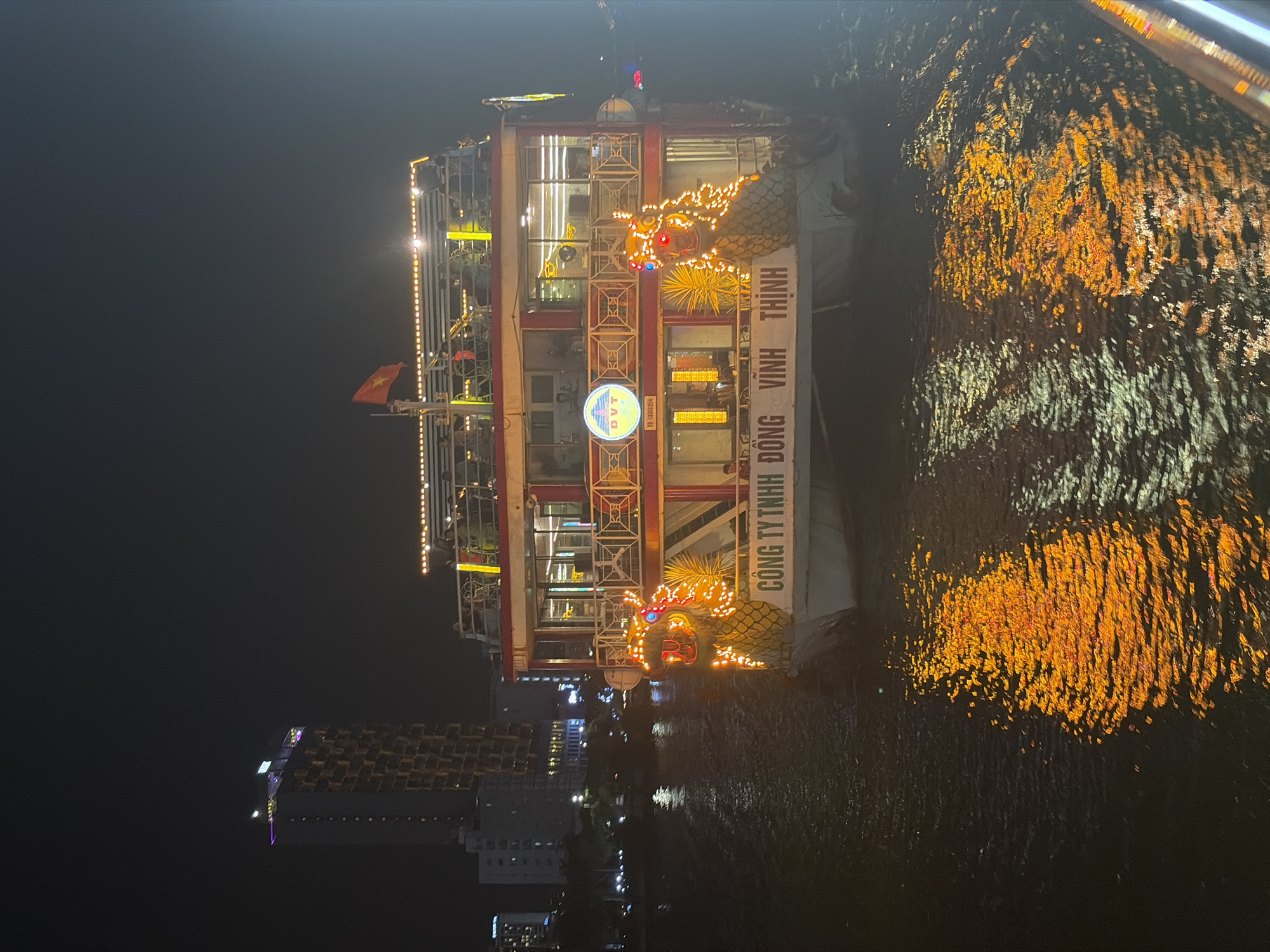 The image, taken at night, captures a brightly illuminated multi-story boat on a body of water, likely a river or bay, in Đà Nẵng, Vietnam. The boat is the central subject, adorned with numerous warm-toned lights, including elaborate, illuminated dragon figures on its side, suggesting a festive or ornamental purpose, perhaps a dinner cruise or floating restaurant. A Vietnamese flag, red with a yellow star, is visible flying from the boat's upper deck.
The water surface reflects the boat's vibrant lights, creating a mosaic of shimmering orange and white hues. In the background, city lights are visible, with a tall, distinctively patterned building standing out on the far left. Other smaller buildings and distant lights dot the cityscape along the water's edge. The overall atmosphere is lively and illuminated against the dark night sky.
Visible text on the side of the boat reads vertically: "CÔNG TY TNHH ĐỒNG VĨNH THỊNH" (Dong Vinh Thinh Co., Ltd.). Below a circular logo, the characters "DVT" and a number "VP-3100154" are also discernable. The image is rotated 90 degrees counter-clockwise.