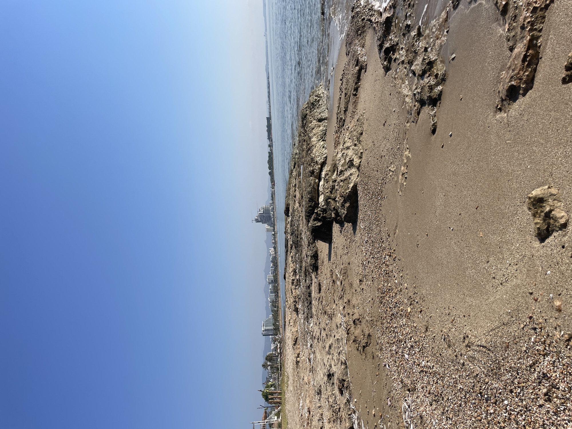 A wide shot captures a beach scene with a clear blue sky overhead. The foreground is dominated by sandy and rocky terrain, with scattered pebbles and debris. In the mid-ground, the sea meets the shore, with gentle waves lapping against the rocks. Further in the distance, a coastal city skyline is visible, characterized by numerous buildings, including hotels and apartments. Palm trees and other vegetation can be seen along the coastline. The overall environment suggests a sunny day at a beach resort.