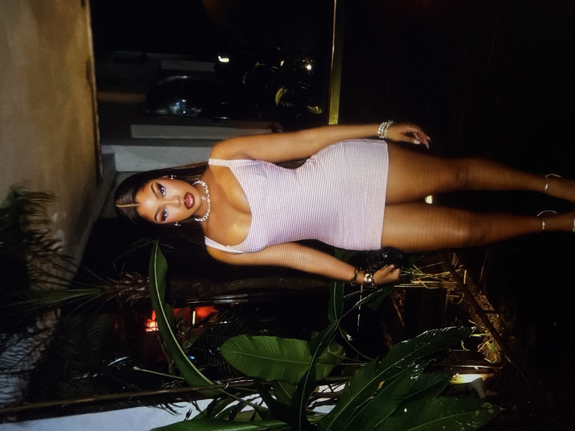 In a dimly lit outdoor setting, a Black woman stands and poses for the camera. She is wearing a shimmering pink, form-fitting mini dress, a diamond choker necklace, and bracelets on both wrists. Her makeup is bold, featuring dramatic winged eyeliner and a deep red lipstick. Her long, dark hair is parted in the middle and styled straight. She is positioned in front of lush green tropical plants and what appears to be a building structure, with a dark background suggesting nighttime. The scene is suggestive of a night out in a stylish location, possibly a restaurant or lounge, in Abuja, Nigeria.