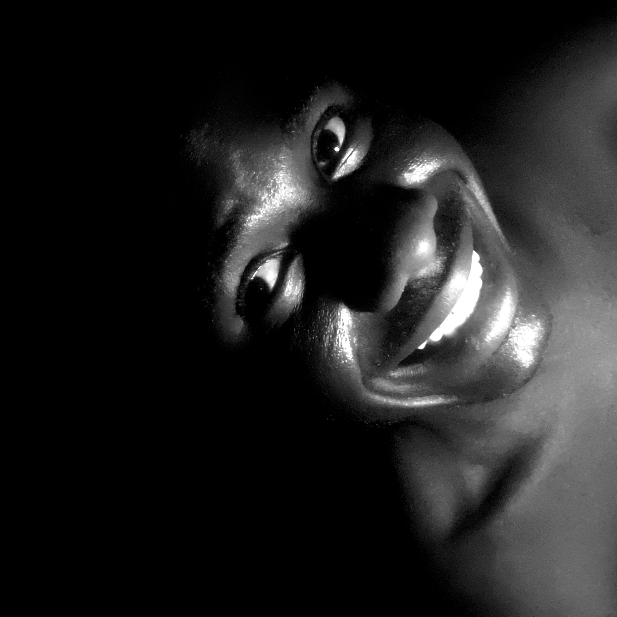 A close-up, black and white portrait shows a person with dark skin smiling broadly, exposing their teeth. The image is characterized by dramatic lighting, with the person's face emerging from deep shadows on the left side of the frame. The skin appears to have a glossy sheen, suggesting moisture or oil. The eyes are directed towards the camera, exhibiting a sense of engagement. The head is tilted at an angle, and the background is entirely dark. There is no indication of the city of Narayi, Nigeria, or any specific location.