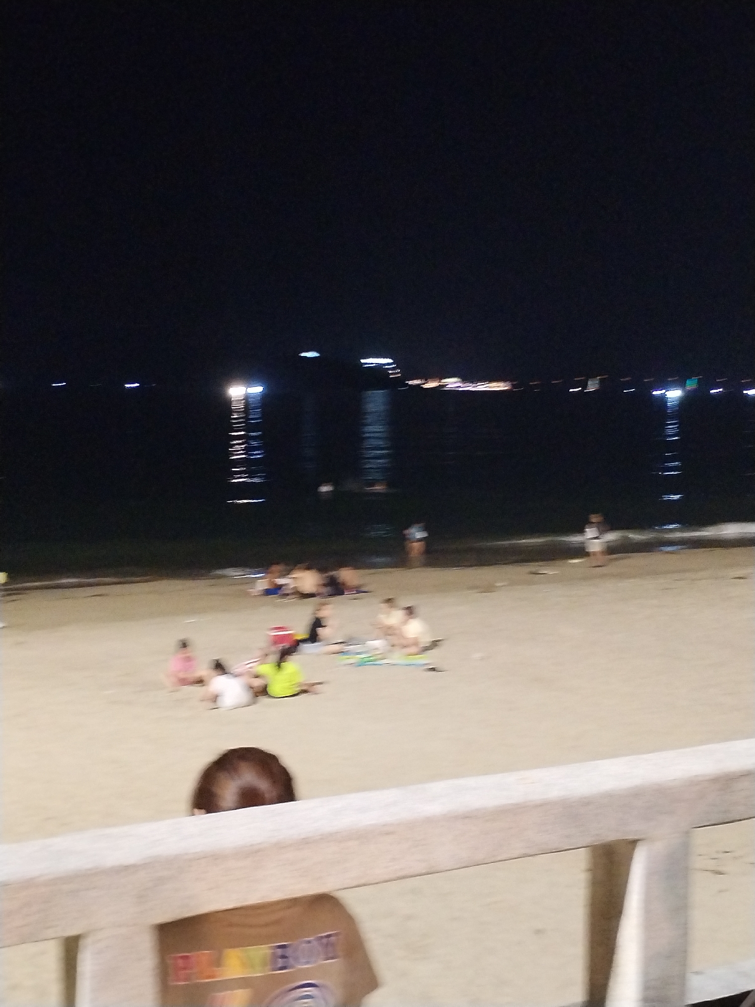 The photo, taken at night in Nha Trang Ward, Vietnam, shows a beach scene. The beach is sandy with several groups of people scattered around. Some people are sitting or lying on the sand. In the background, there is a body of water and distant city lights are visible across the water. A wooden railing is in the foreground, slightly obscuring the view of the beach, and a person's head and shoulders can be seen behind the railing. The person is wearing a shirt with the word "PLAYBOY" on it. The lighting suggests it's nighttime, and the artificial lights in the distance reflect on the water.