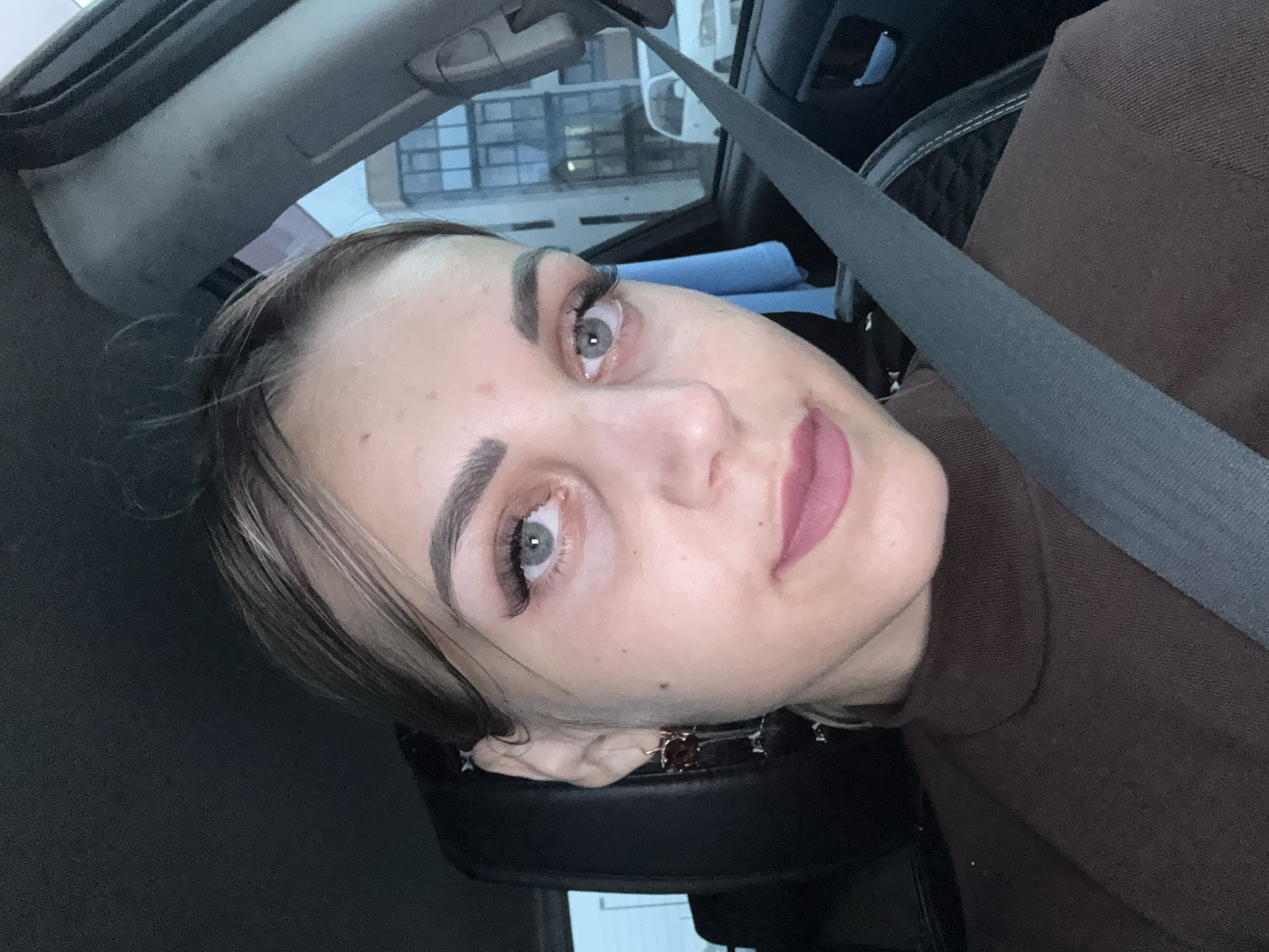 A woman is taking a selfie inside a car, wearing a brown turtleneck sweater and a seatbelt. She has on makeup including false eyelashes and lipstick. She is also wearing a dangling earring on her left ear. The background shows a building with windows and a white car parked outside, suggesting an urban setting. The location is identified as Yuzhno-Sakhalinsk, Russia.