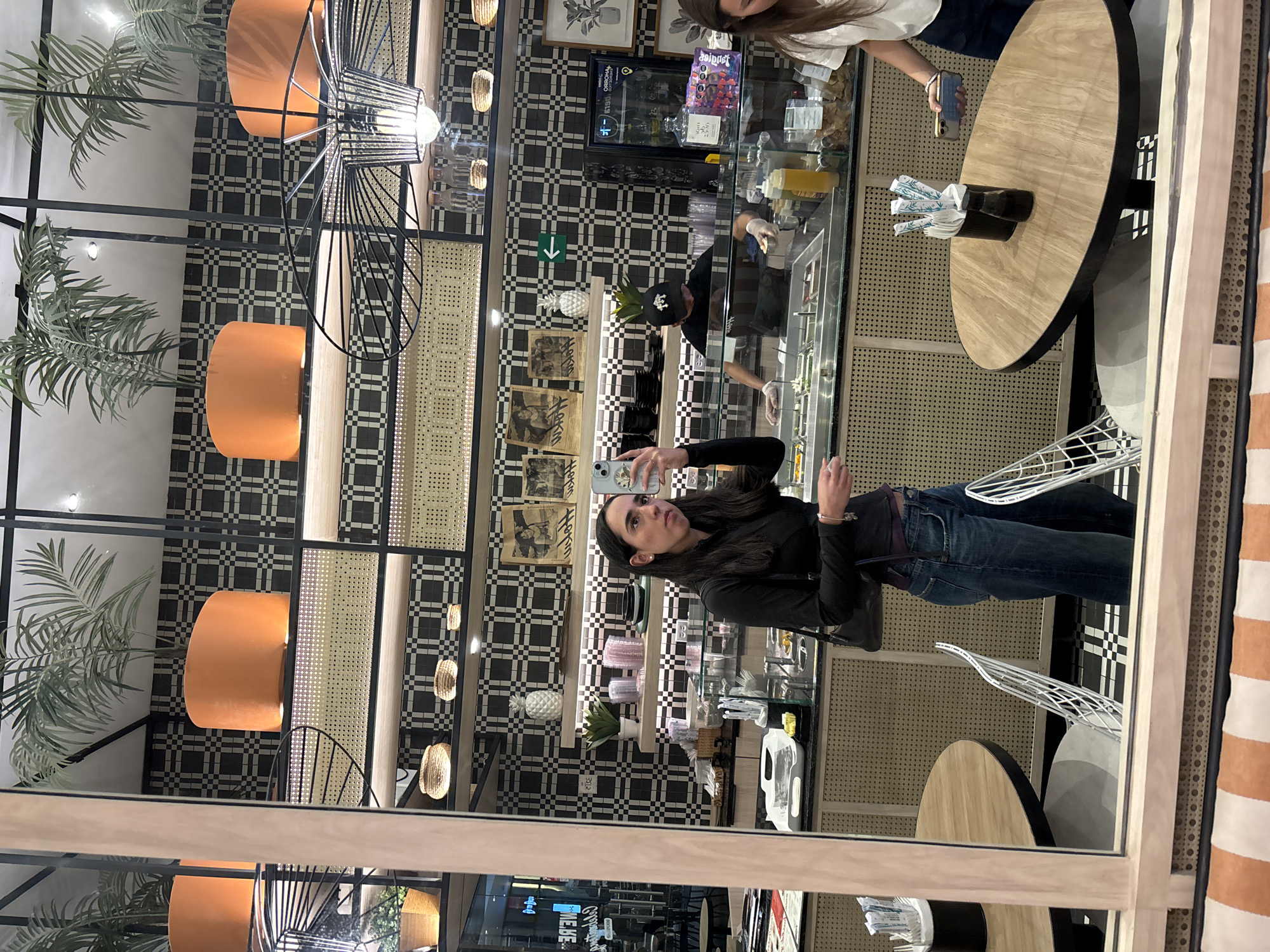 A reflection of a woman taking a photo of a display case in a restaurant is captured in a large mirror. The woman, wearing a black top and blue jeans, is holding a smartphone up to her face. The display case contains food items and is being attended to by a worker wearing gloves and a cap. The restaurant's interior includes black-and-white checkered walls, woven textures, and hanging orange lights with metal frames. Tables with white chairs are visible, and other people are present in the background. The location appears to be in a city; the name of the city is not clear from the visual information available.