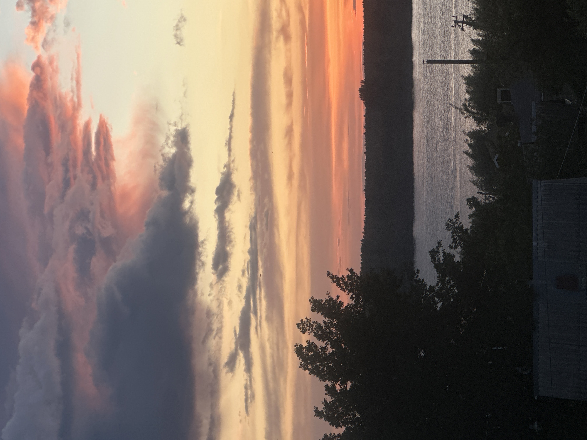 The image shows a landscape at dusk, possibly in Chaikovsky, Russia. To the right, there's a body of water (likely a river or lake) with a line of trees or forest on the opposite bank. Utility poles are visible near the water's edge. In the foreground, a silhouette of trees obscures the view. The sky displays a sunset with streaks of orange, yellow, and gray clouds. To the left, there are darker, more defined cloud formations with shades of gray and purple. A building is seen partially obscured by the trees on the bottom right corner.