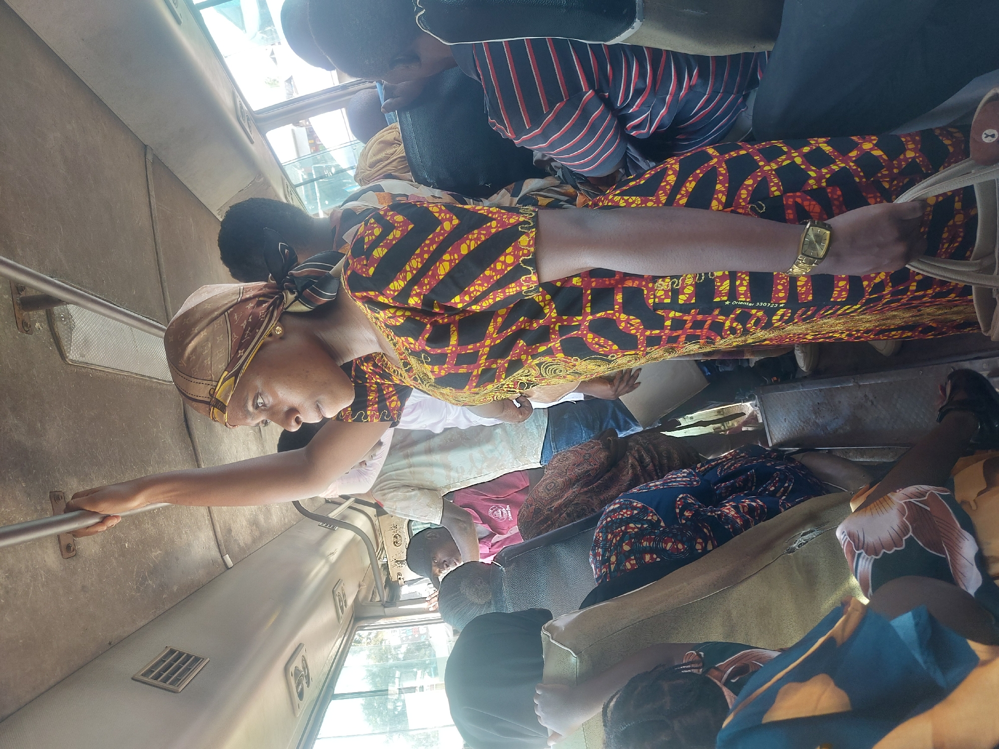 The image depicts the crowded interior of a public transport vehicle, likely a bus or 'daladala,' in Dar es-Salaam, Tanzania. The scene is captured during daylight hours, with bright natural light entering through the windows.
Numerous passengers fill the vehicle, some standing and holding onto silver grab bars, while others are seated on beige-colored upholstery. In the foreground, a woman stands prominently, holding a horizontal silver bar for stability. She wears a striking patterned dress in black, yellow, and red geometric designs, complemented by a coordinating headscarf. A gold-colored wristwatch is visible on her left wrist. On her dress, near the waist, the text "Orientar 330723" can be partially seen, suggesting a fabric brand or code. Her expression is calm and attentive.
Behind her, other passengers are visible. One individual is seen from the back, wearing a black and red striped shirt. Further inside the vehicle, additional passengers, many dressed in similarly colorful and patterned attire, are either standing or seated. The setting reflects a typical busy commuting environment within an urban area.